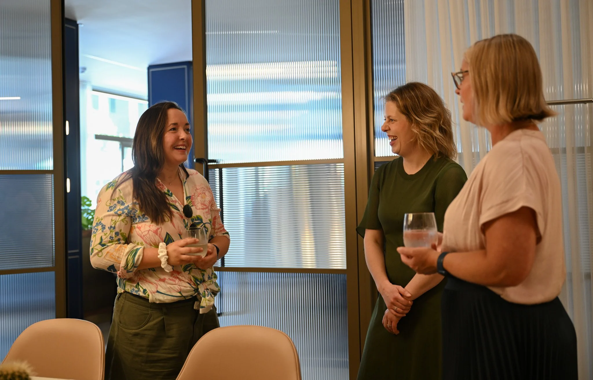Three women standing and shared laughing in a modern room, each holding a drink, engaged in conversation.