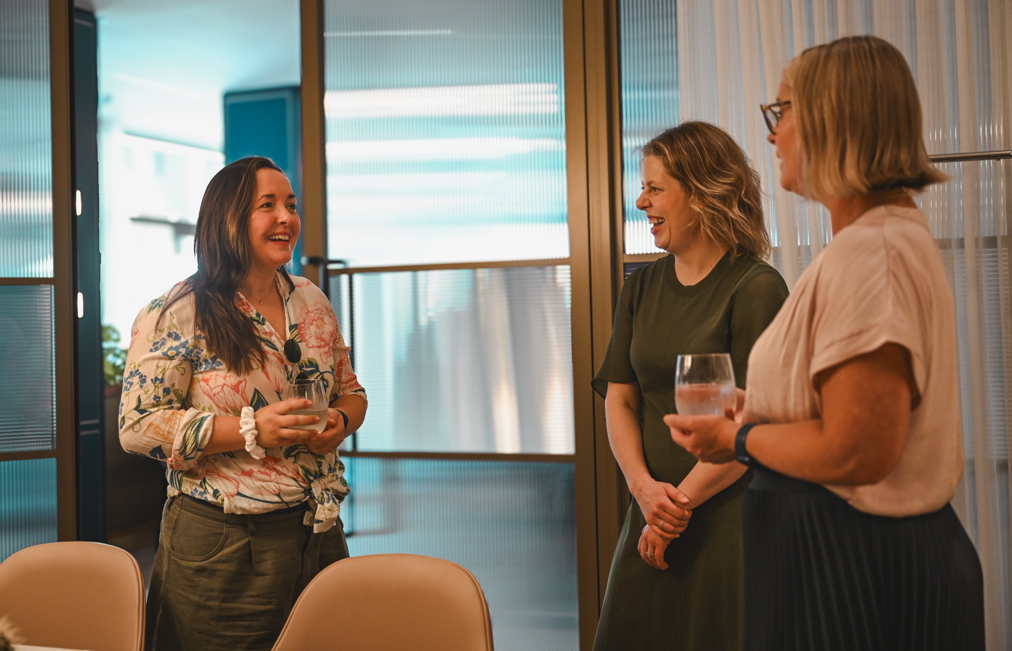 Three women are standing and talking with drinks in their hands in a room with modern decor and glass doors in the background.