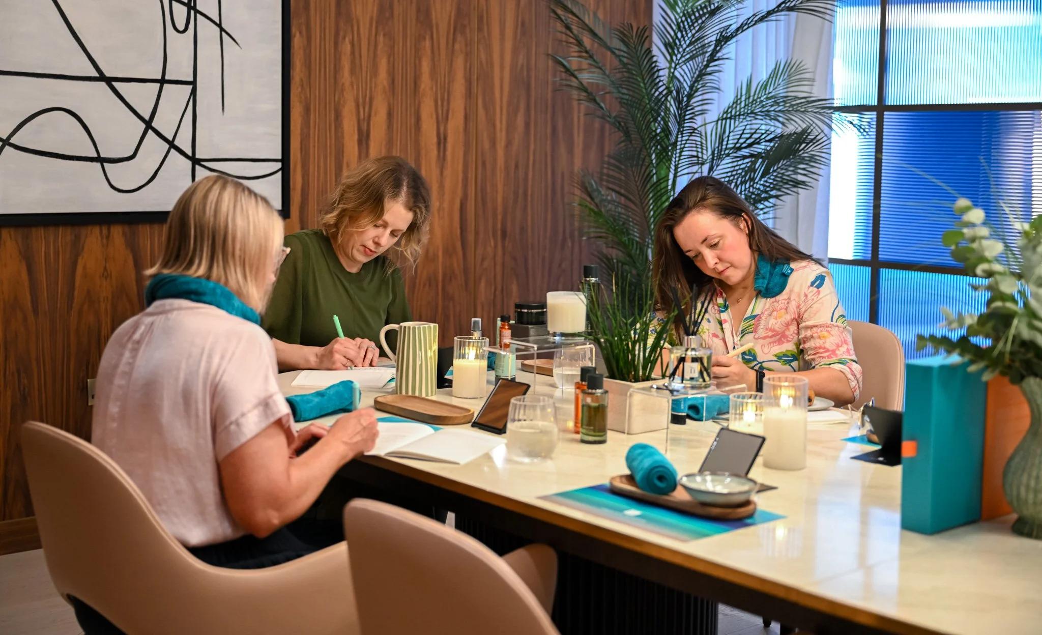 Four women sit at a table working on a project, with notebooks, pens, and various supplies on the table. The room has wooden walls, a large abstract artwork, and a large window with blinds, decorated with potted plants.