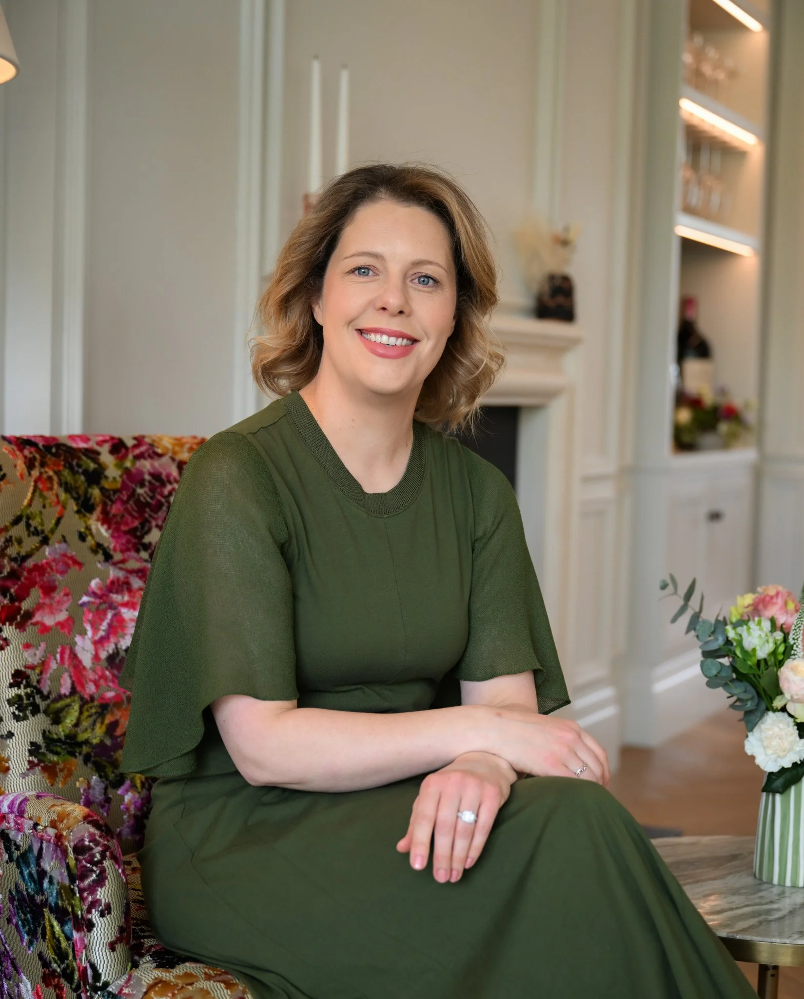 A woman with shoulder-length light brown hair and blue eyes sitting in a living room, wearing a green dress, smiling at the camera, with flowers in a vase nearby.