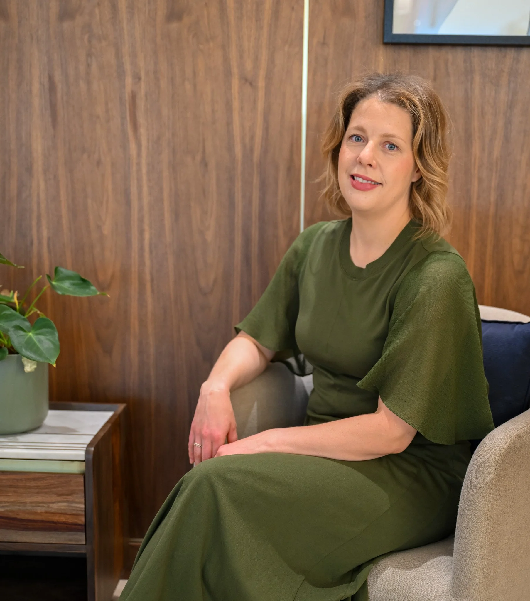A woman with shoulder-length curly hair and blue eyes, wearing a green top, sitting indoors against a wooden wall with a framed picture visible. There is a potted plant to her left.