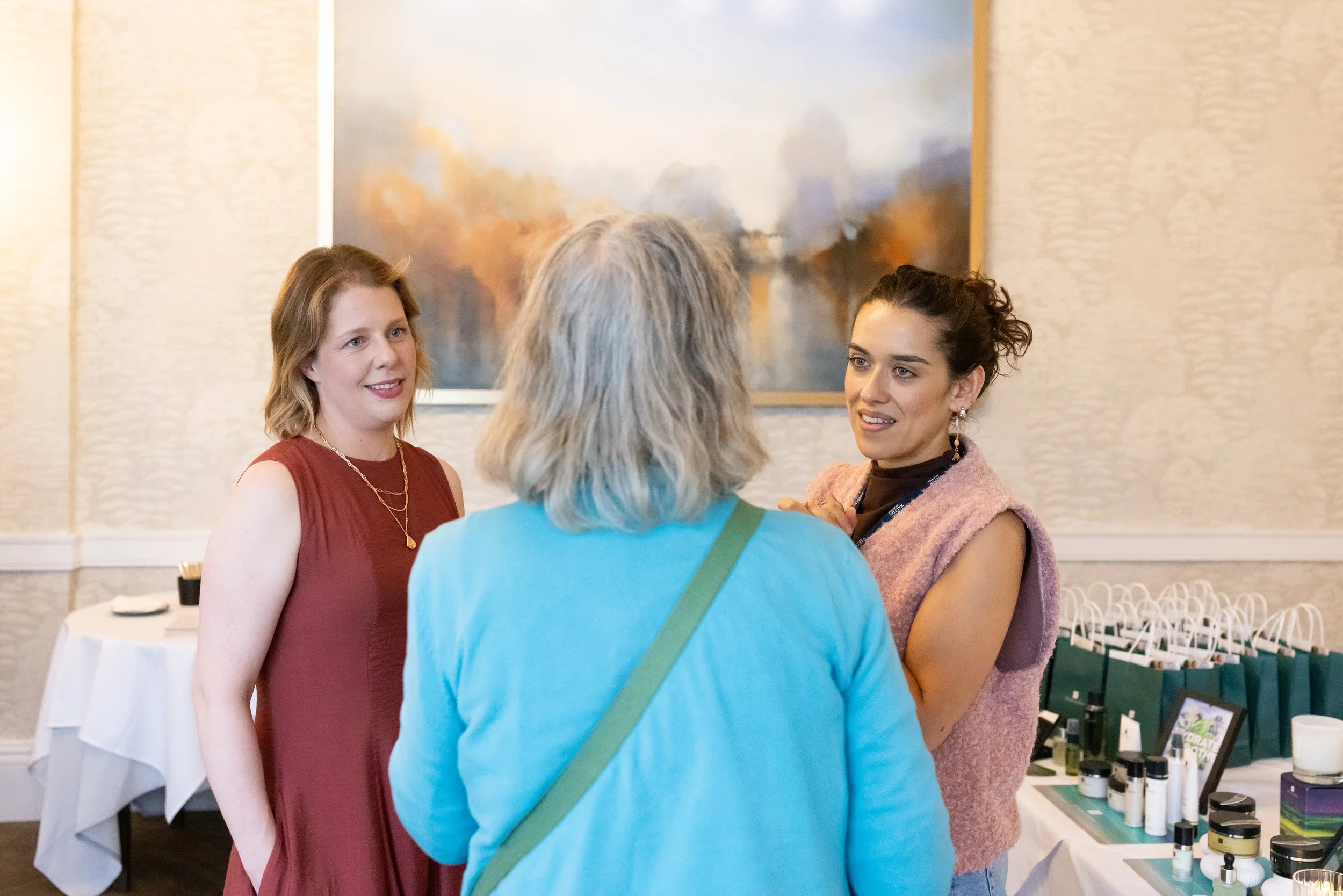 Three women engaged in conversation at an event, with a large abstract painting on the wall behind them and a table with skincare or cosmetic products to the right.