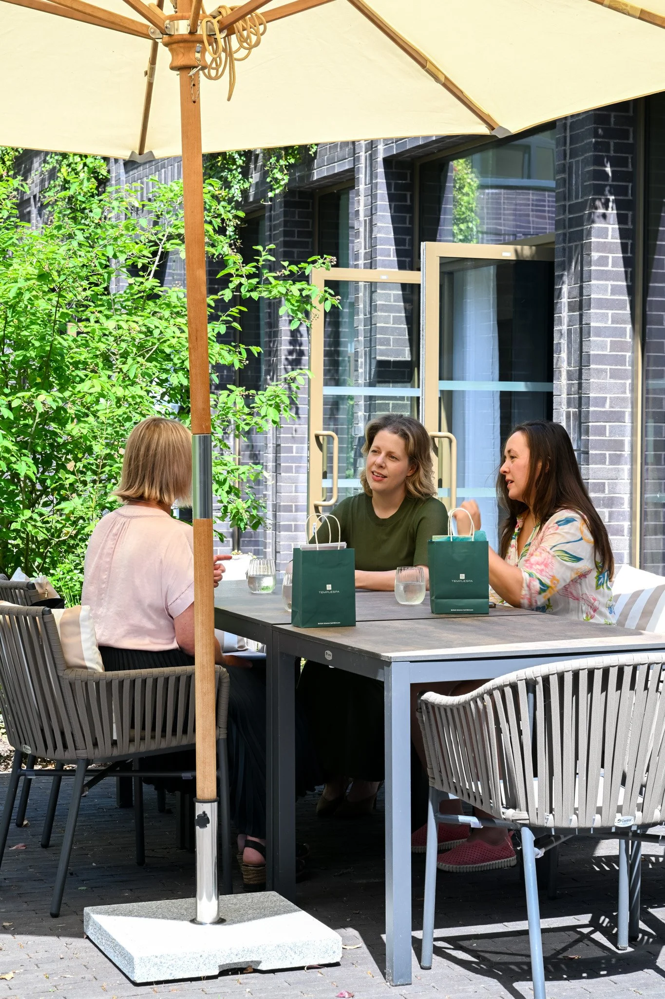 Four women are sitting around an outdoor dining table, engaged in conversation. The table has several green shopping bags and glasses of water. A large beige patio umbrella shades the area, and there are green bushes and a modern dark brick building in the background.