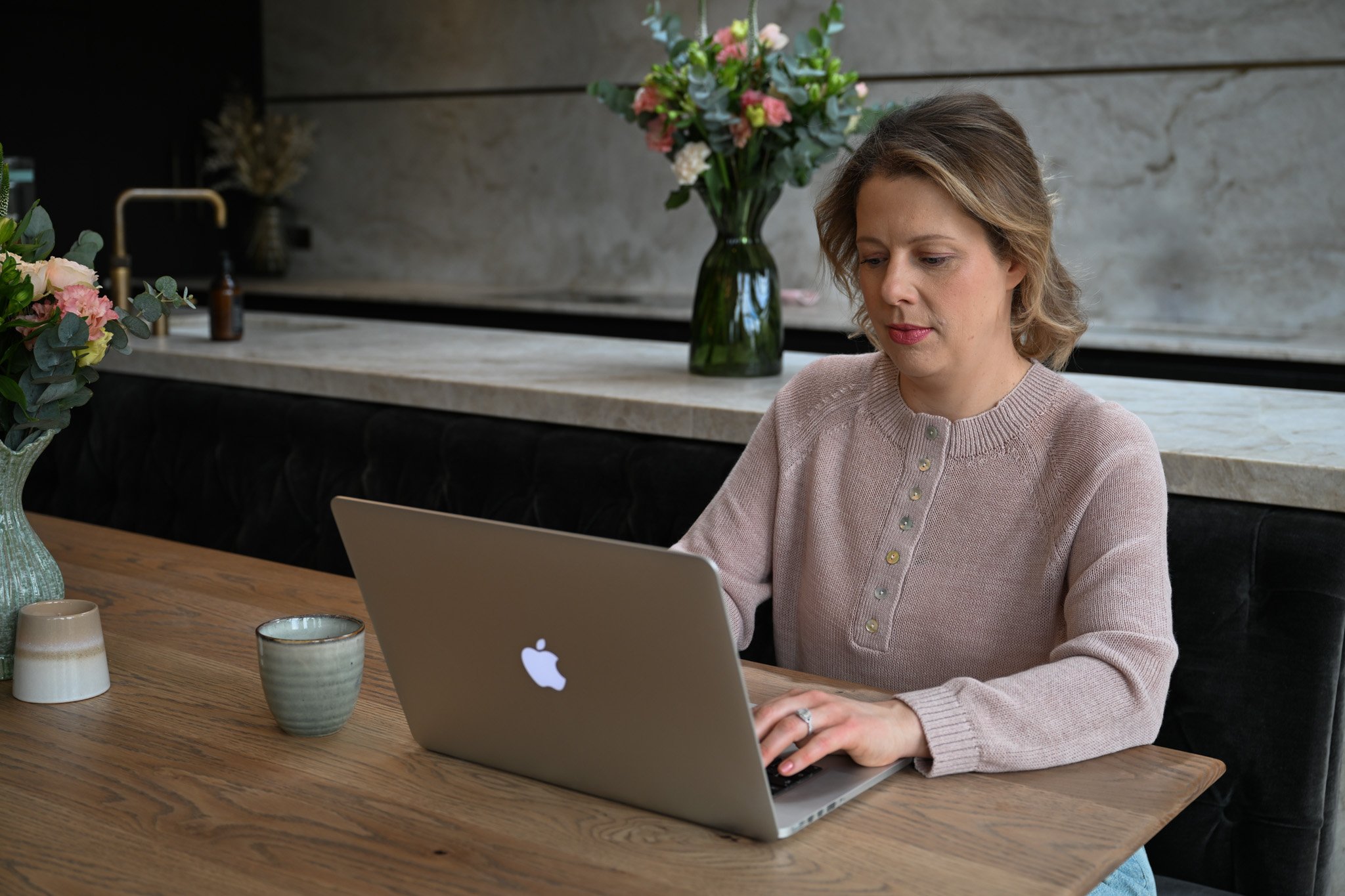 A woman with short blonde hair in a pink knit sweater working on a silver MacBook laptop at a wooden table, with a large bouquet of pink and white flowers in a green vase behind her in a modern kitchen.