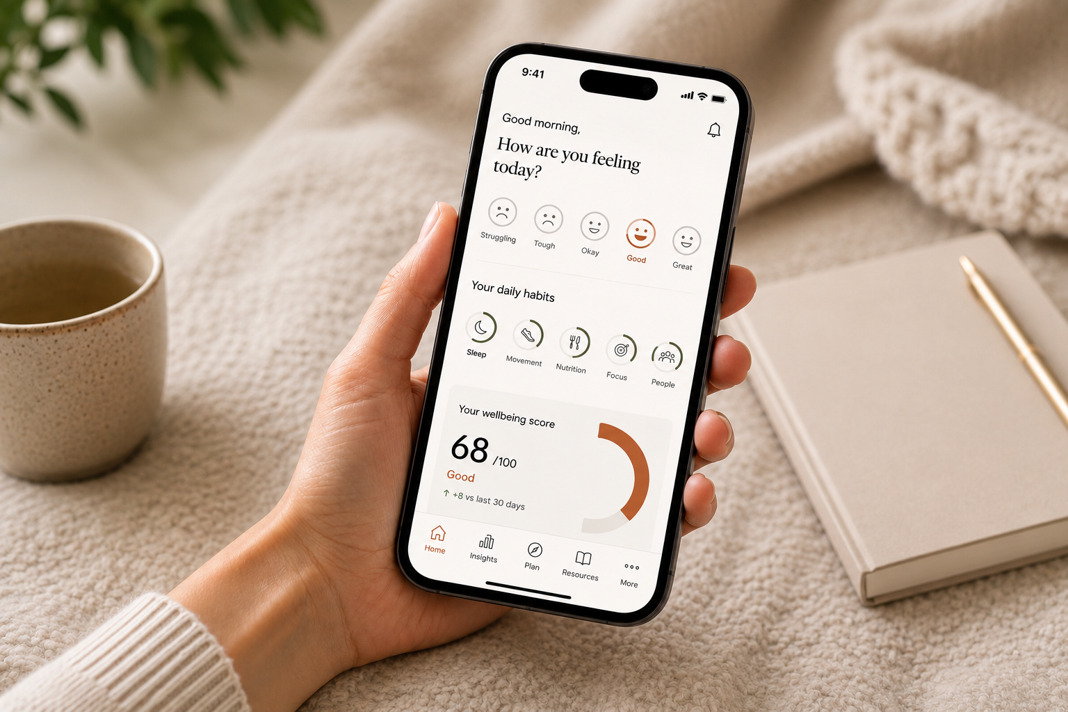 A person holding a smartphone displaying a wellness app with mood, daily habits, and wellbeing score, with a coffee cup and a notebook on a cozy beige blanket nearby.