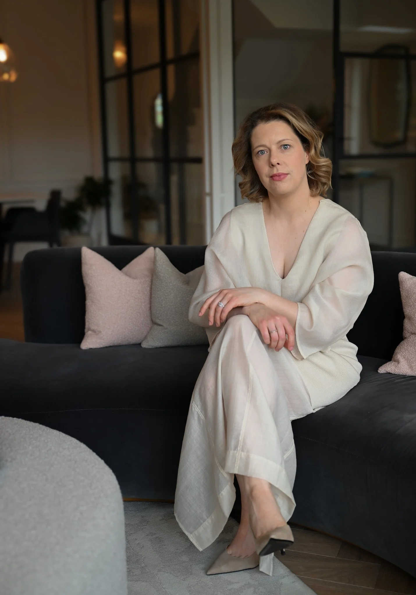 A woman sitting on a black sofa in a modern, stylish living room, wearing a cream-colored dress, with her legs crossed and hands resting on her lap, surrounded by light-colored pillows.