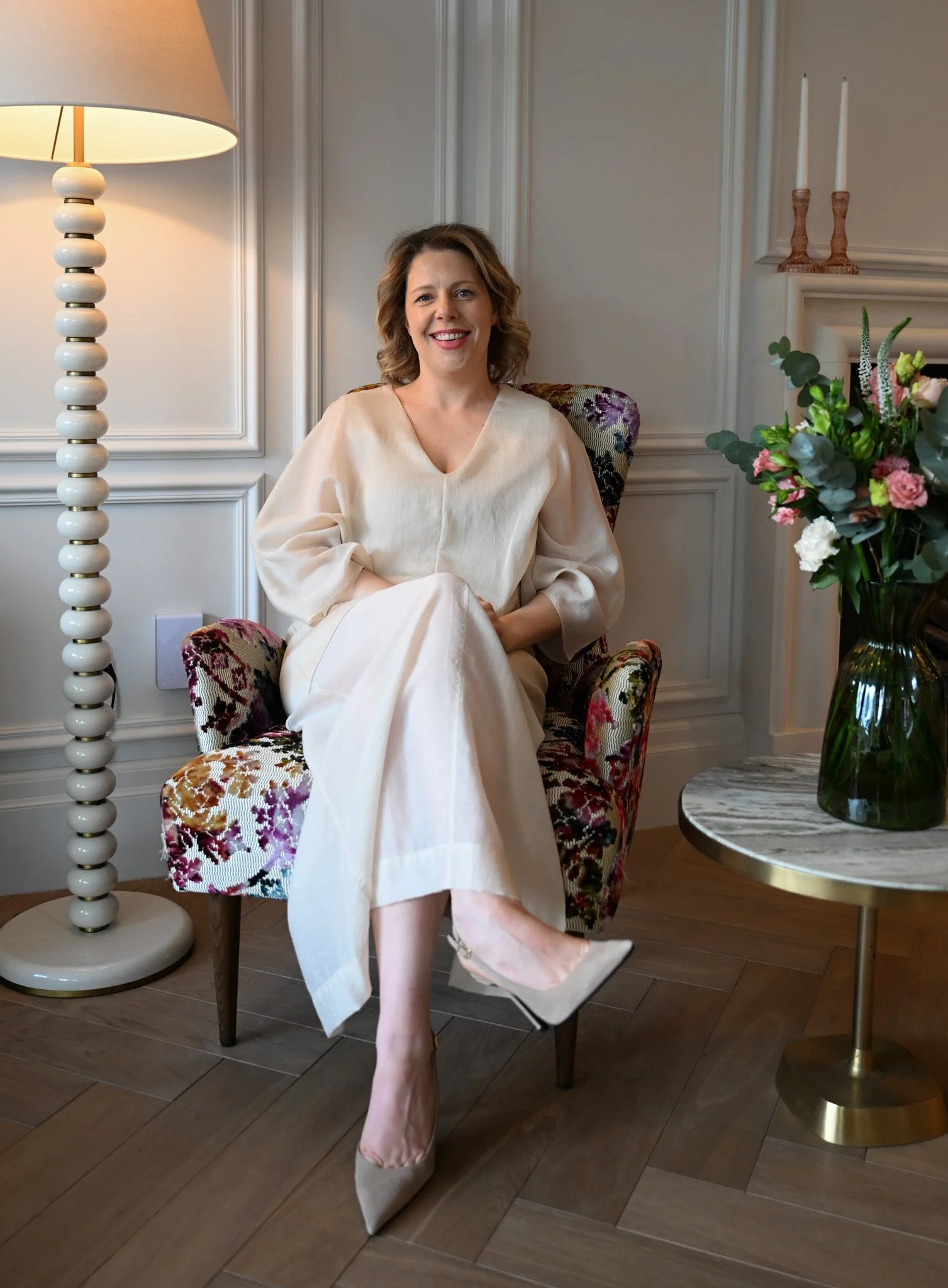 A woman sitting in a floral upholstered armchair, smiling, wearing a cream-colored dress and beige high heels, with a table holding a large green vase with pink and white flowers, and a white lamp with a round base beside her.