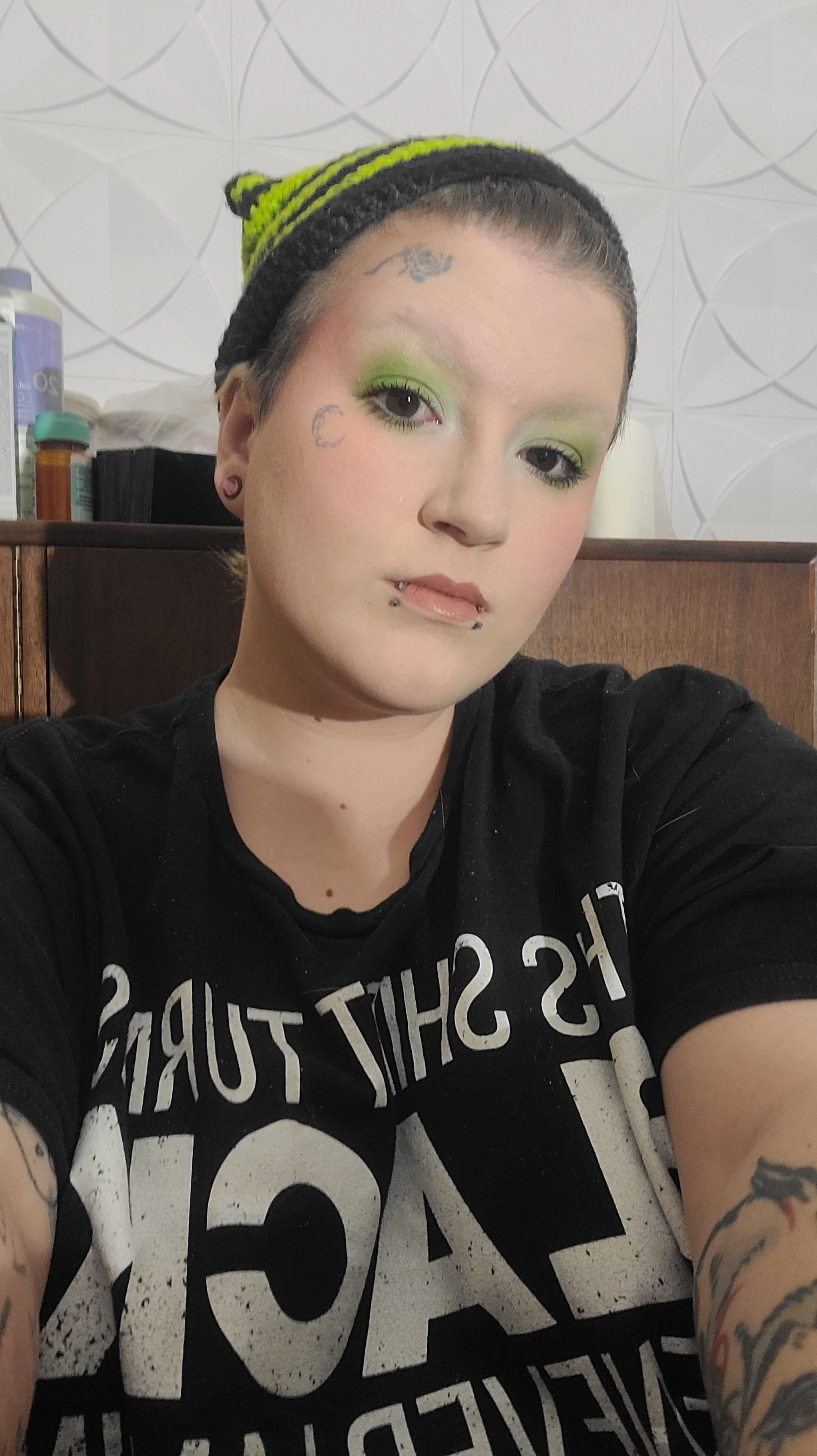 A woman with short dark hair and tattoos on her arms, wearing bright green eyeshadow, a black beanie with green stripes, and a black shirt displaying white text, taking a selfie indoors with a wooden surface and items on it in the background.