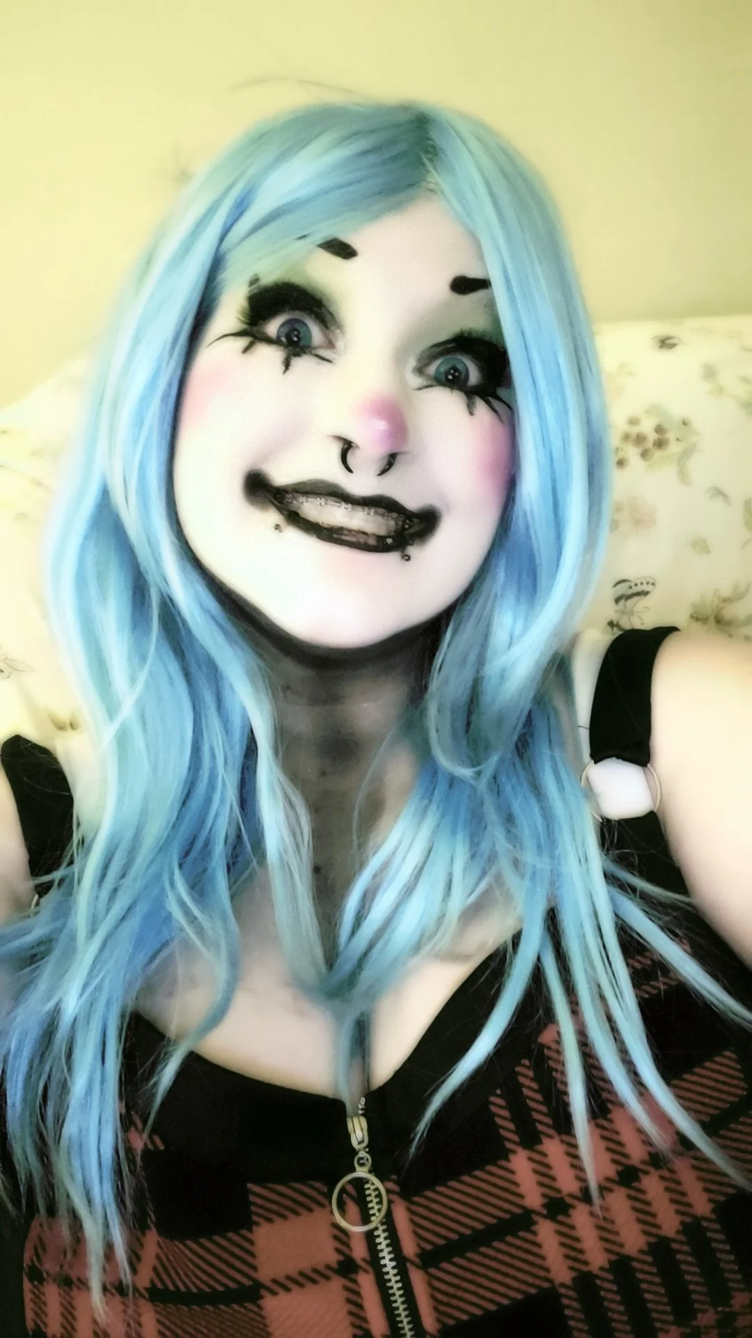 A woman with long, light blue hair and bold makeup, including black eyeliner, long lashes, a pink nose, and black lipstick, taking a selfie indoors.