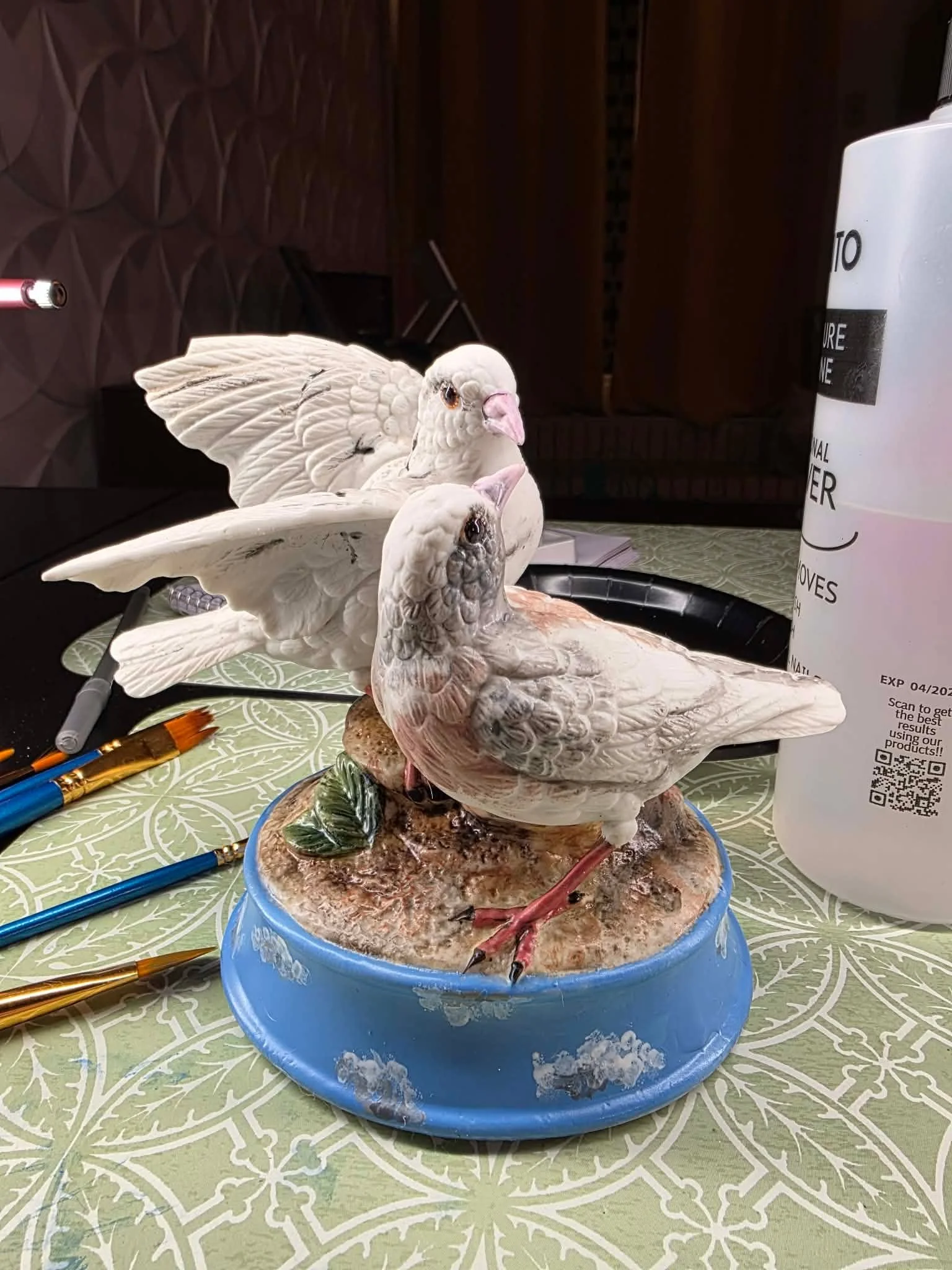 A ceramic sculpture of two white doves with detailed feathers, standing on a textured base with leaves, placed on a patterned green tablecloth. In the background, there are art supplies and a white container.