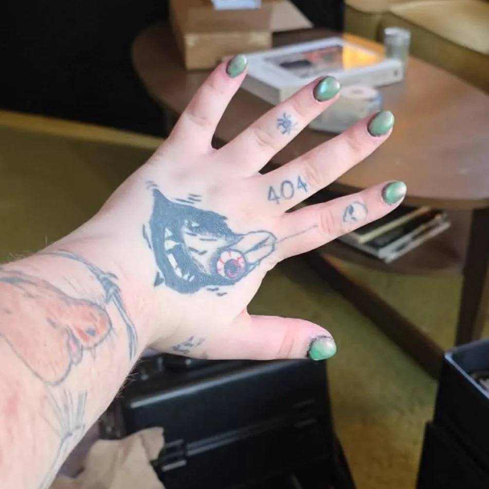 A person's hand with green nail polish showing tattoos. The background includes a wooden table with magazines and a wooden box.