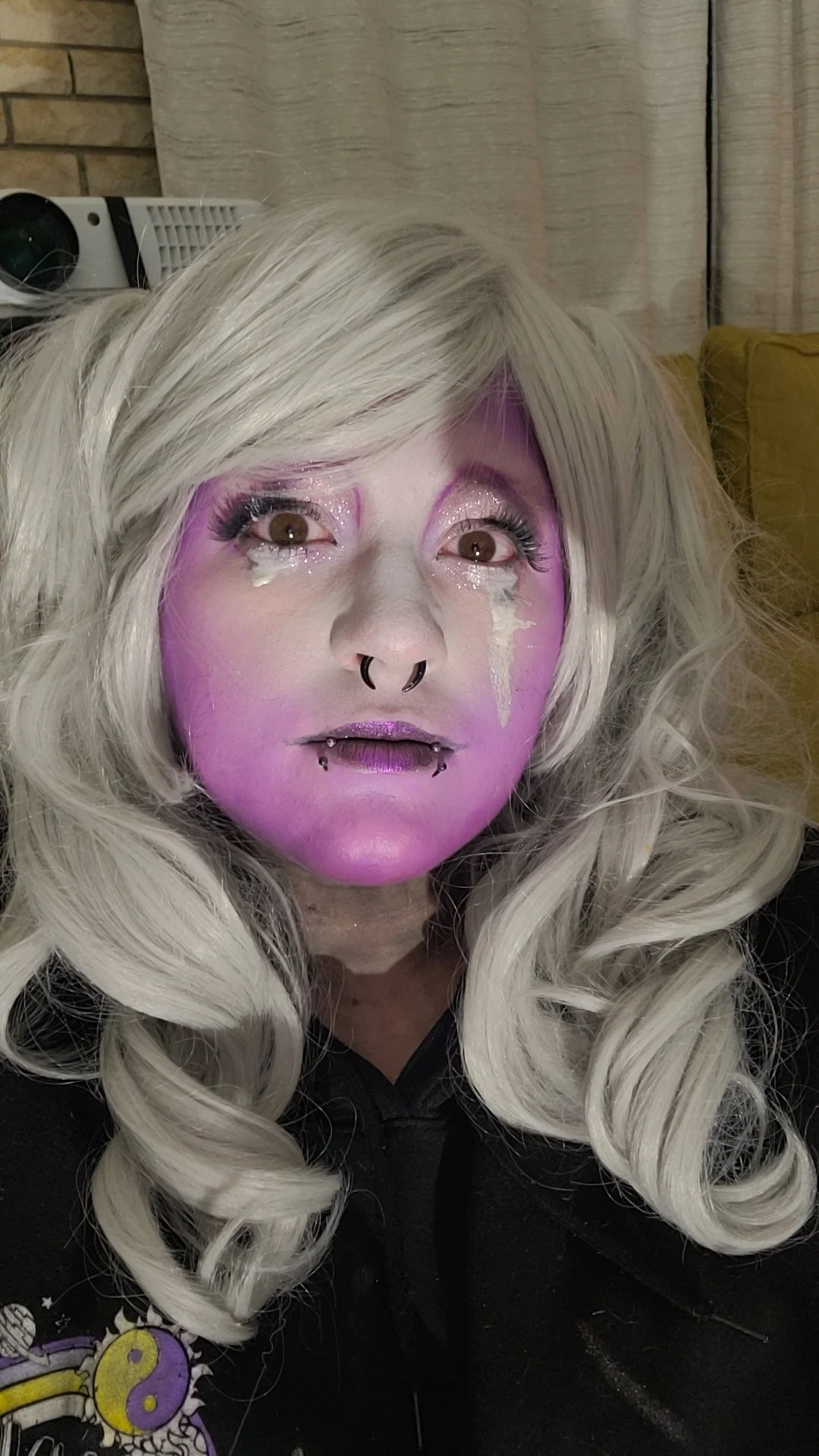 A person with long, curly platinum blonde hair, purple makeup with glitter and drips, and multiple facial piercings, looking directly at the camera.