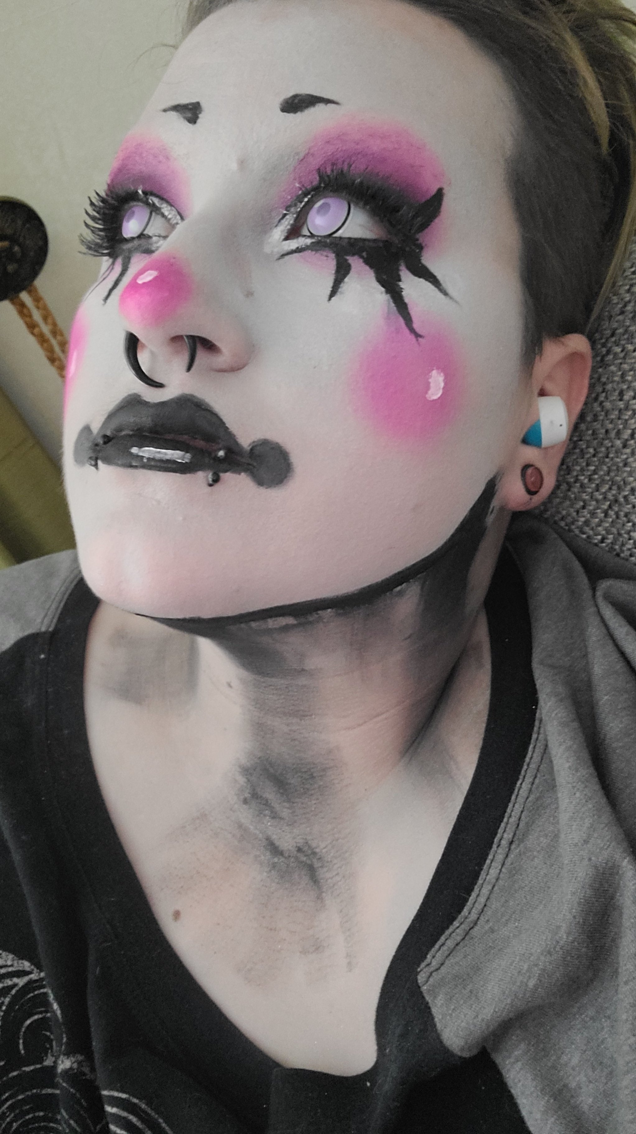 A person with theatrical makeup resembling a clown or mime, featuring exaggerated black eyebrows, pink blush, black lips, and black tears under the eyes, along with purple contact lenses, a septum piercing, and earrings.