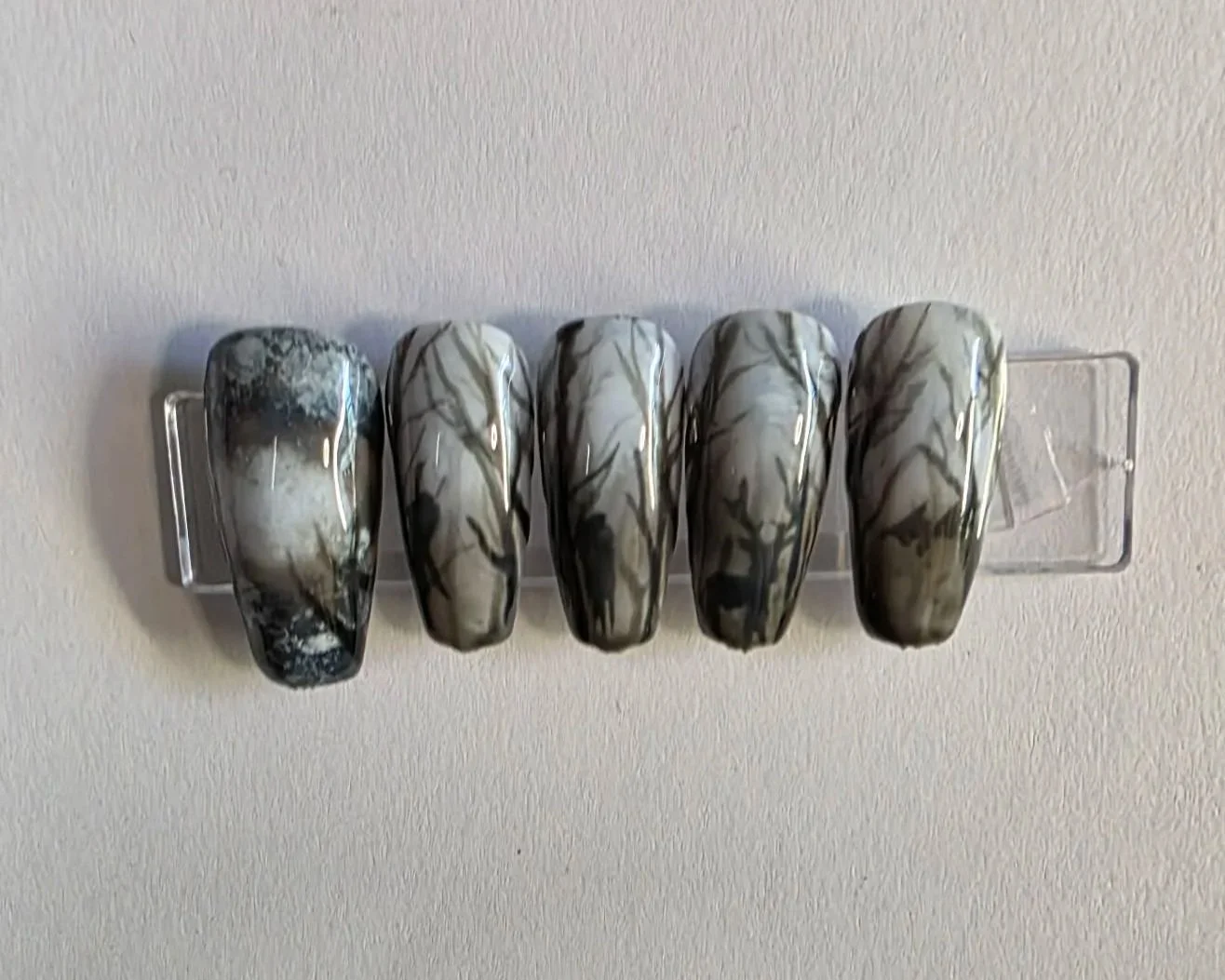 Five painted nails with a black and beige marble pattern displayed on a nail rack.