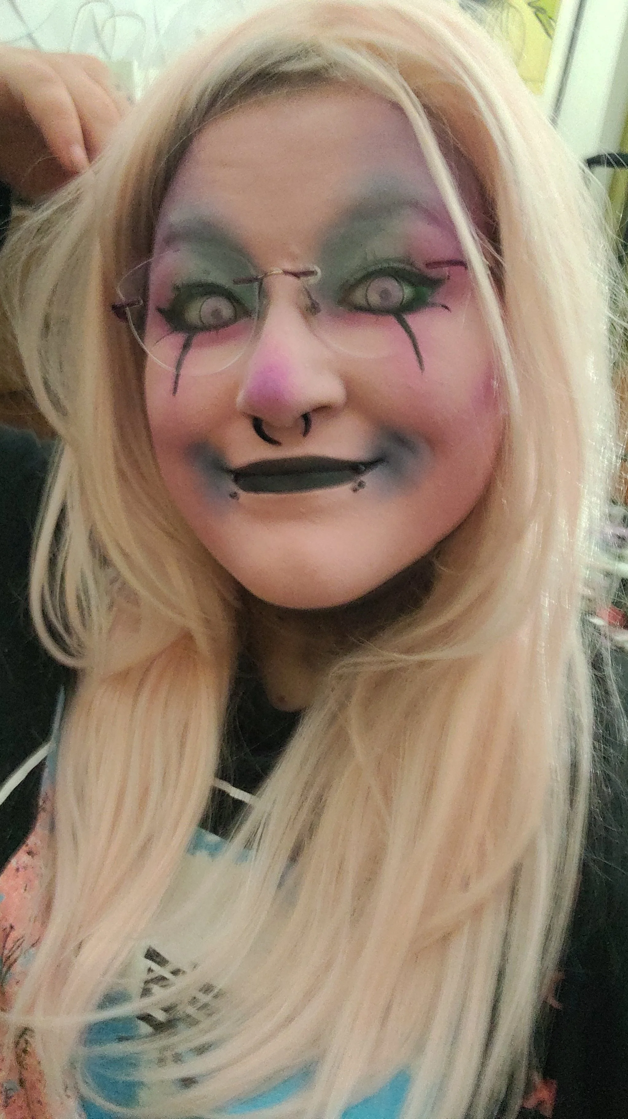 Person with long platinum blonde hair, wearing glasses with artistic makeup featuring pastel colors and black designs, smiling with a septum piercing and lip piercings.