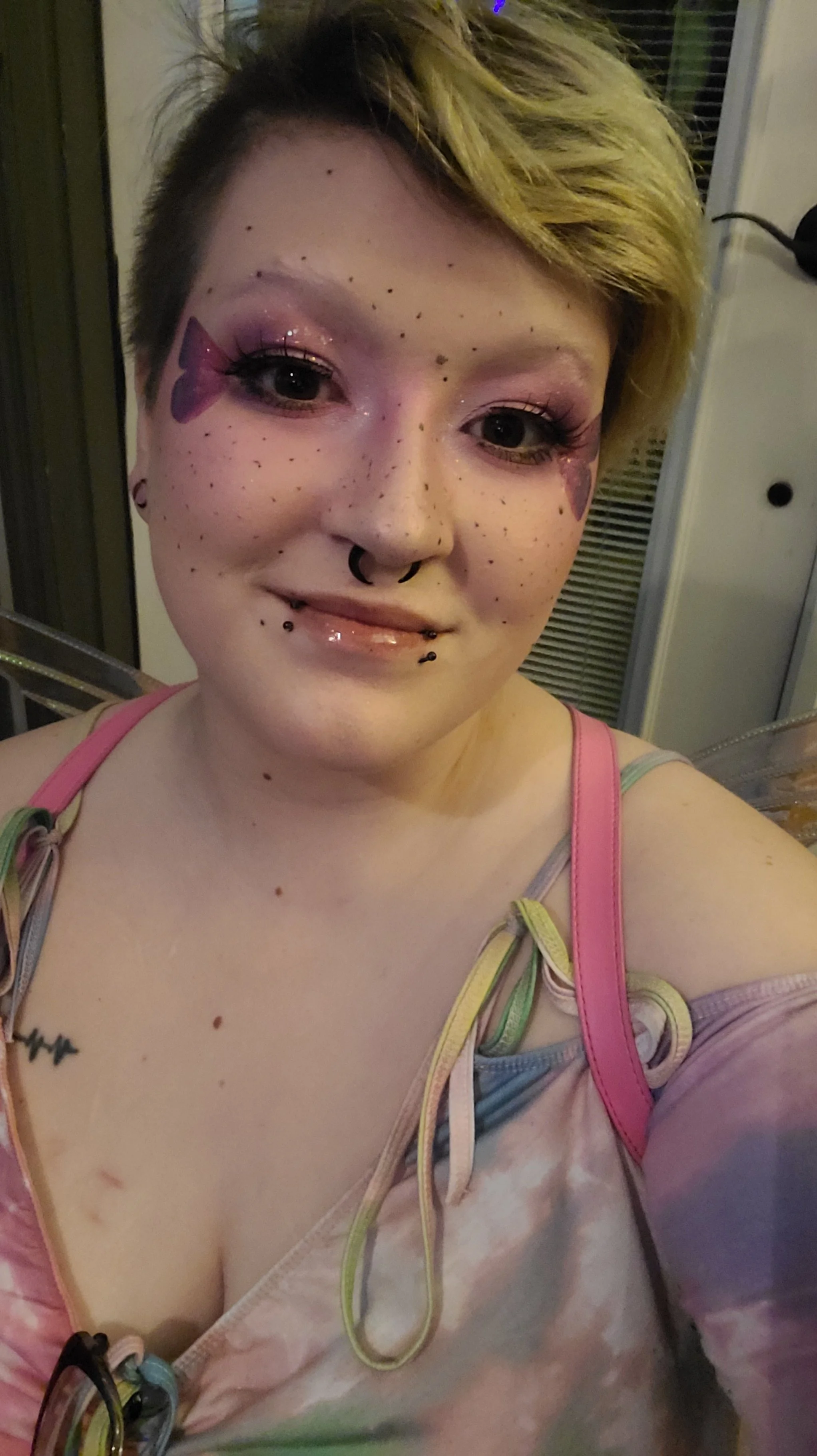 A person with short, light-colored hair and multiple facial piercings, including septum and lip piercings, taking a selfie. They have pink and purple makeup with butterfly designs near their eyes and are wearing a colorful tie-dye top with ribbon det