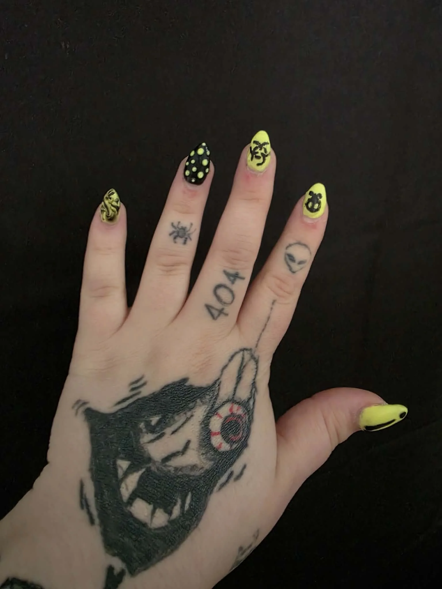 A person's hand with colorful tattoos and bright yellow and black nail art featuring playful designs and patterns.