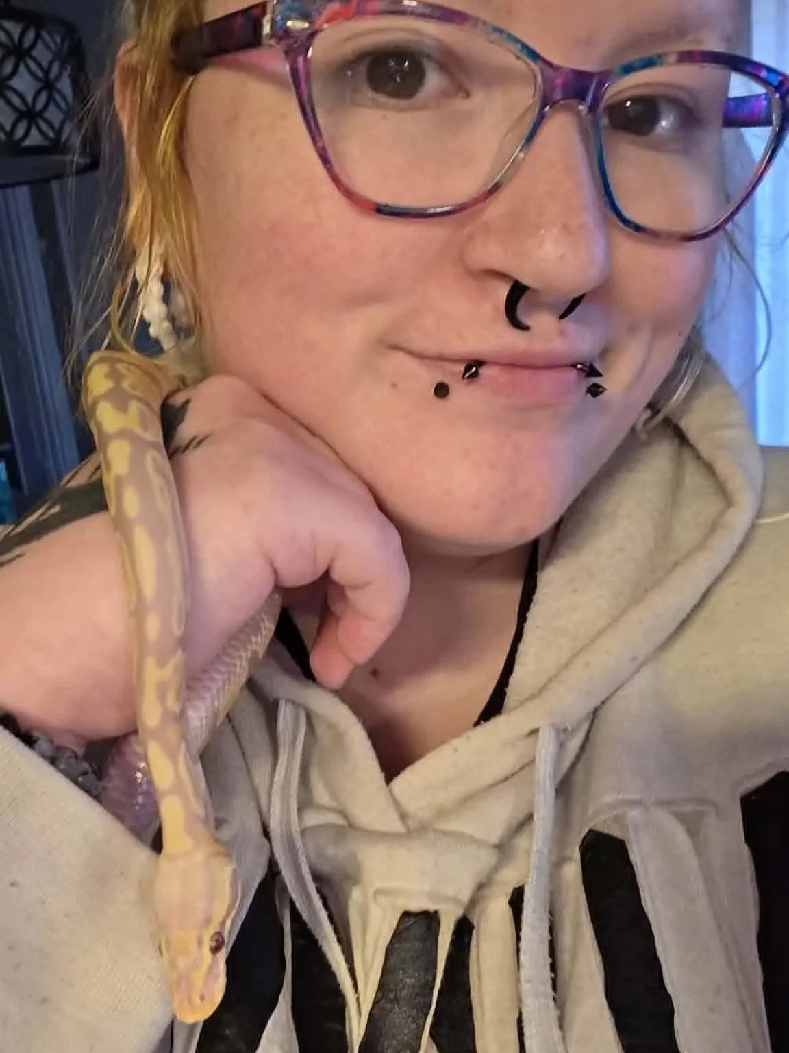 A person wearing colorful glasses and a hoodie with a snake on their hand, smiling.