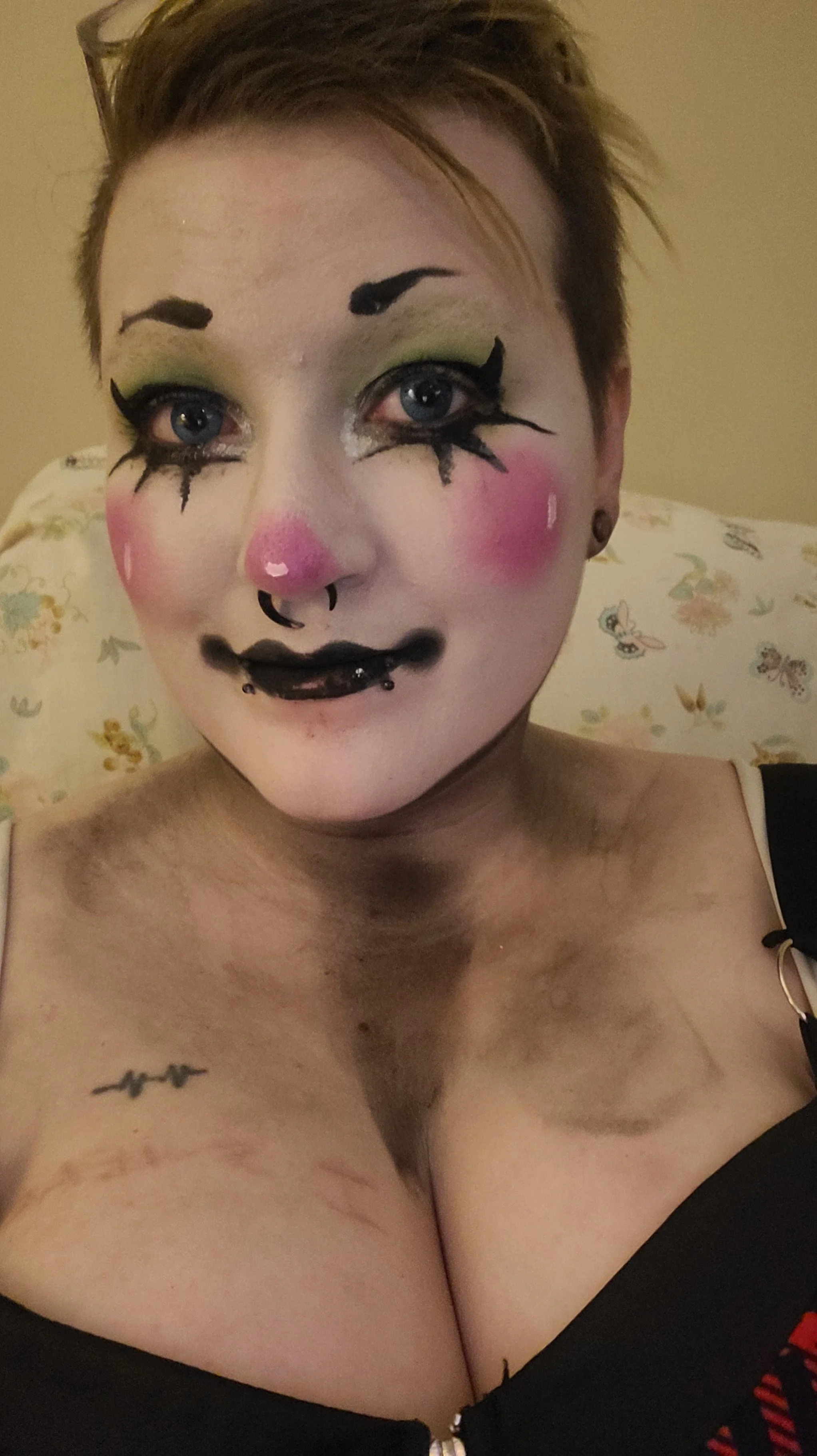 A woman with short hair and blue eyes wearing artistic makeup resembling a clown, including colorful face paint, exaggerated eye makeup, and facial piercings in a selfie shot.