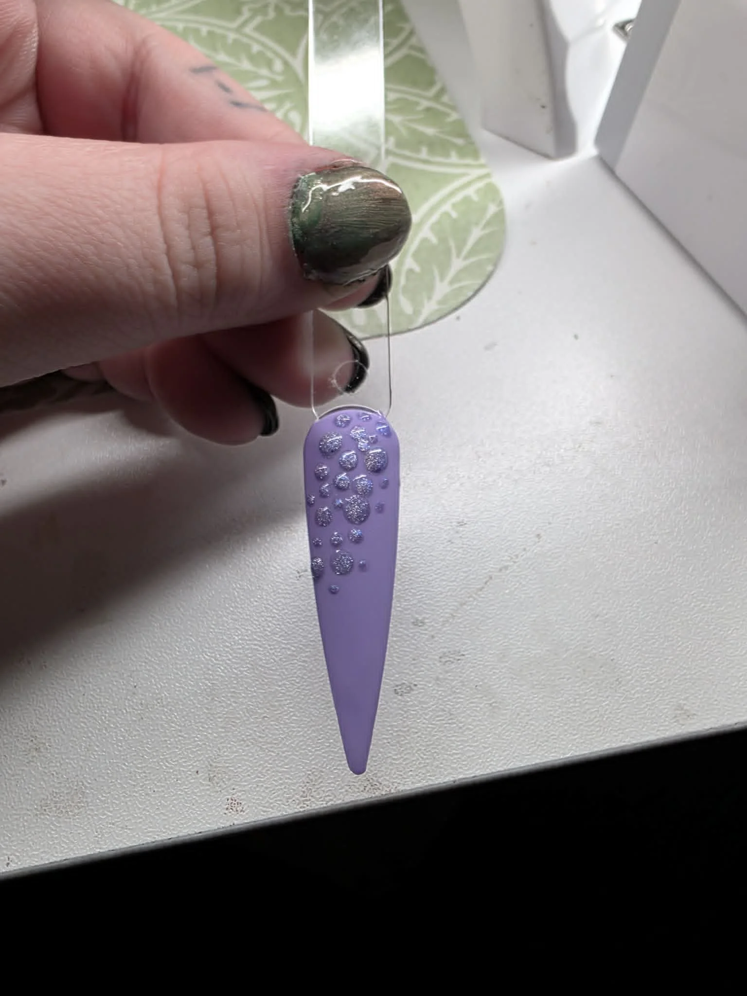 A person's hand with black painted nails holding a purple nail polish strip with rhinestone decorations, suspending in front of a patterned paper and a white surface, with a window in the background.