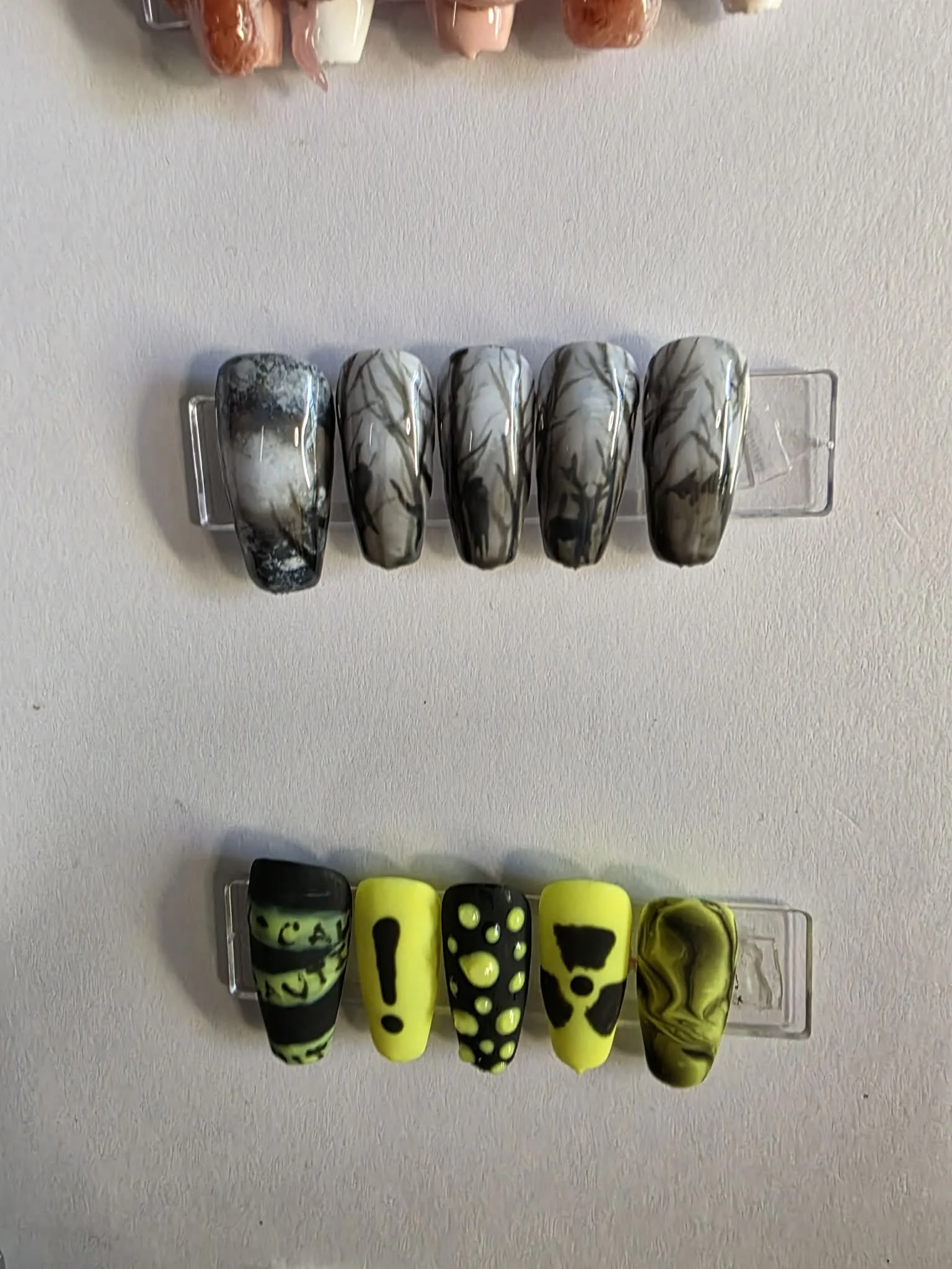 A display of two sets of decorative glass nails, with the top set featuring marble-like gray and black patterns and the bottom set featuring bright yellow backgrounds with black and yellow abstract designs, arranged on clear plastic stands.