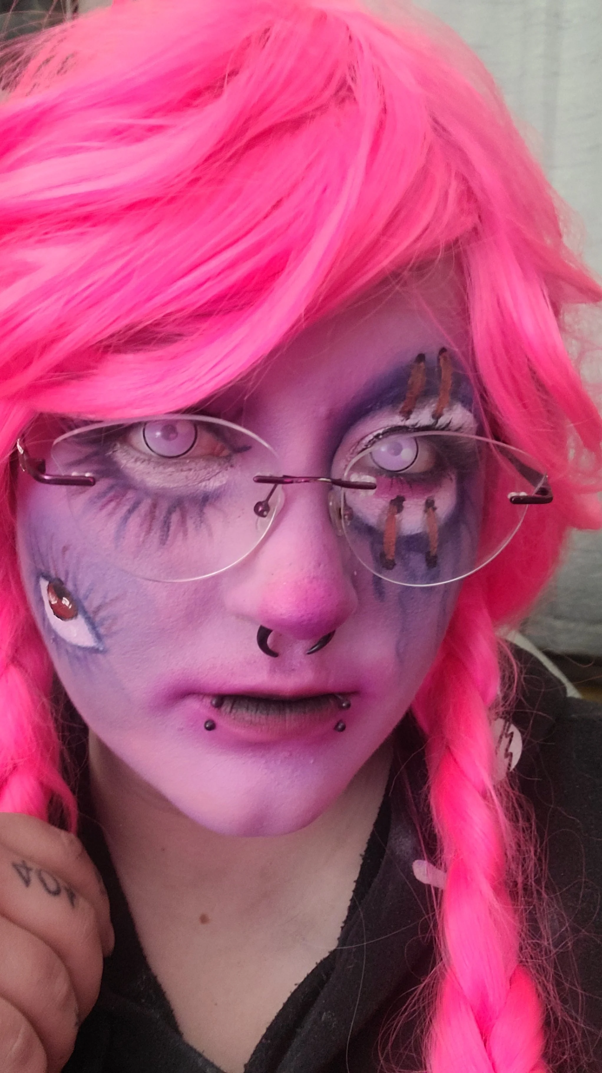 Person with bright pink hair, wearing clear glasses, with face makeup featuring purple and black makeup and small face tattoos, multiple facial piercings, including a septum and lip piercings, and a black shirt.