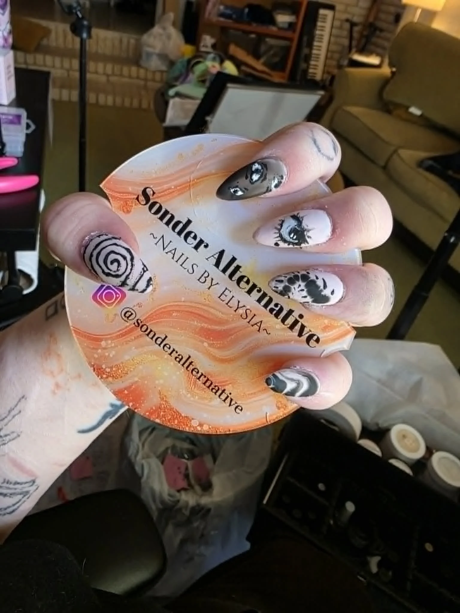 Close-up of a hand with detailed eye and swirl swirl nail art holding a container of Sutton's by Elixir adhesive with a background showing a cluttered living room with a brick wall, couch, bookshelf, and musical keyboard.