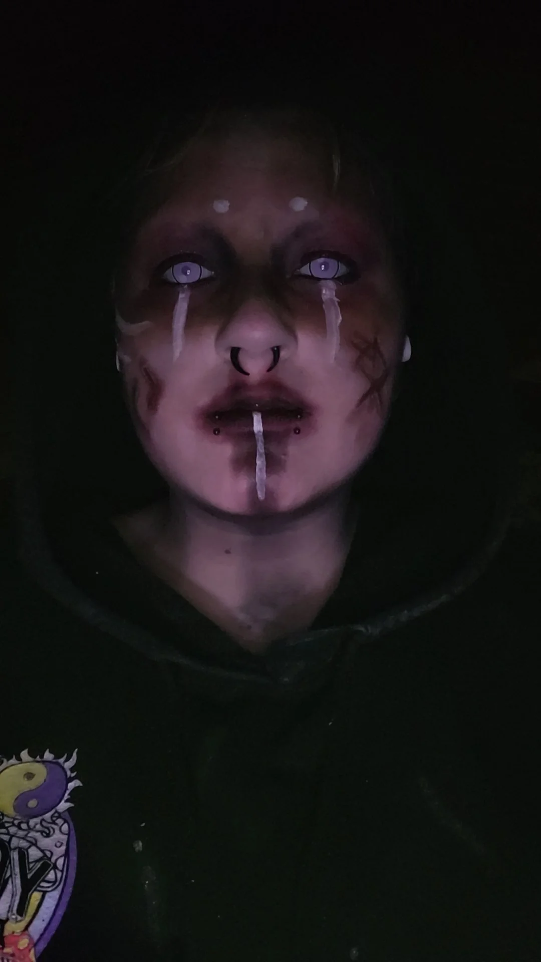 A person with pale skin and facial piercings looking directly at the camera, with makeup or face paint resembling tears and scars, wearing a black hoodie and a shirt with a colorful cartoon graphic.