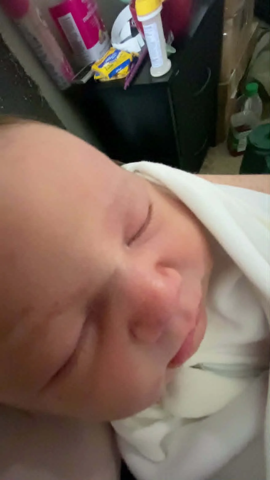 Close-up of a sleeping child's face with closed eyes, partially covered with a white blanket, in a cluttered room with pantry items and cleaning supplies in the background.