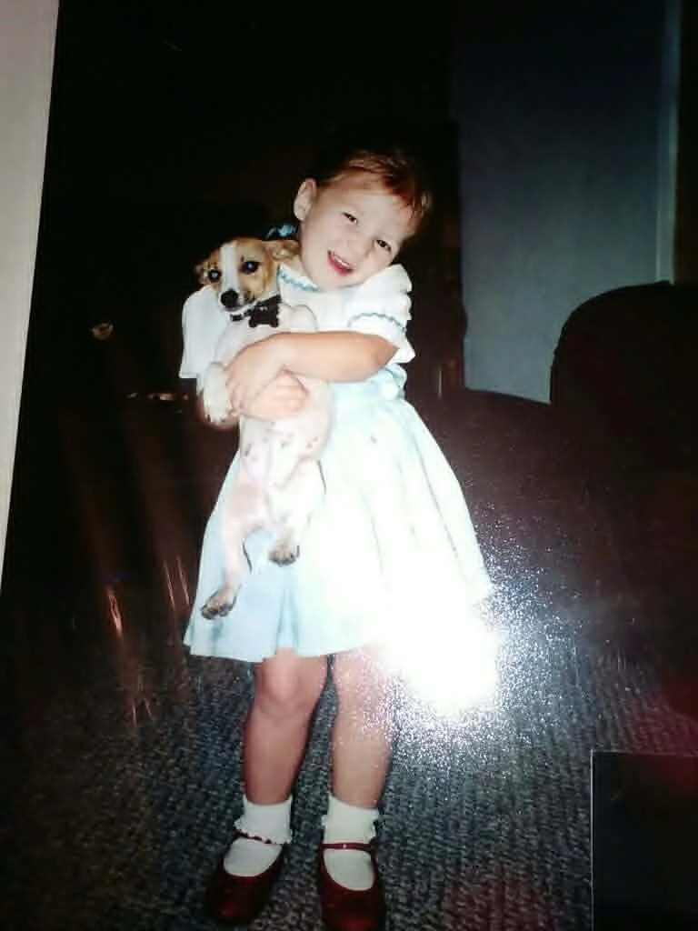 A young girl in a white dress holding a small dog with a collar, standing on a carpeted floor inside a room.