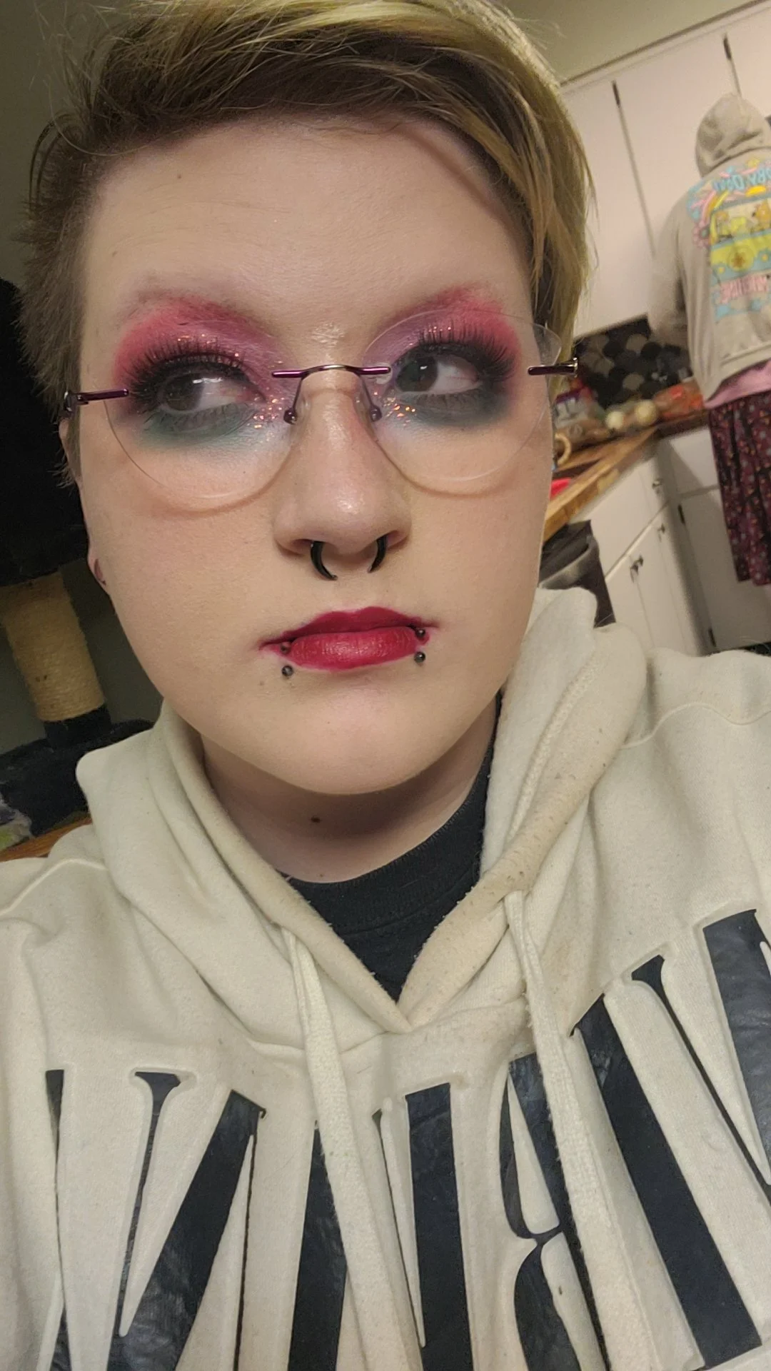 A person with short hair wearing glasses, makeup with pink and purple eyeshadow, red lipstick, and multiple facial piercings, including septum and lip piercings, taking a selfie in a room that appears to be a kitchen.
