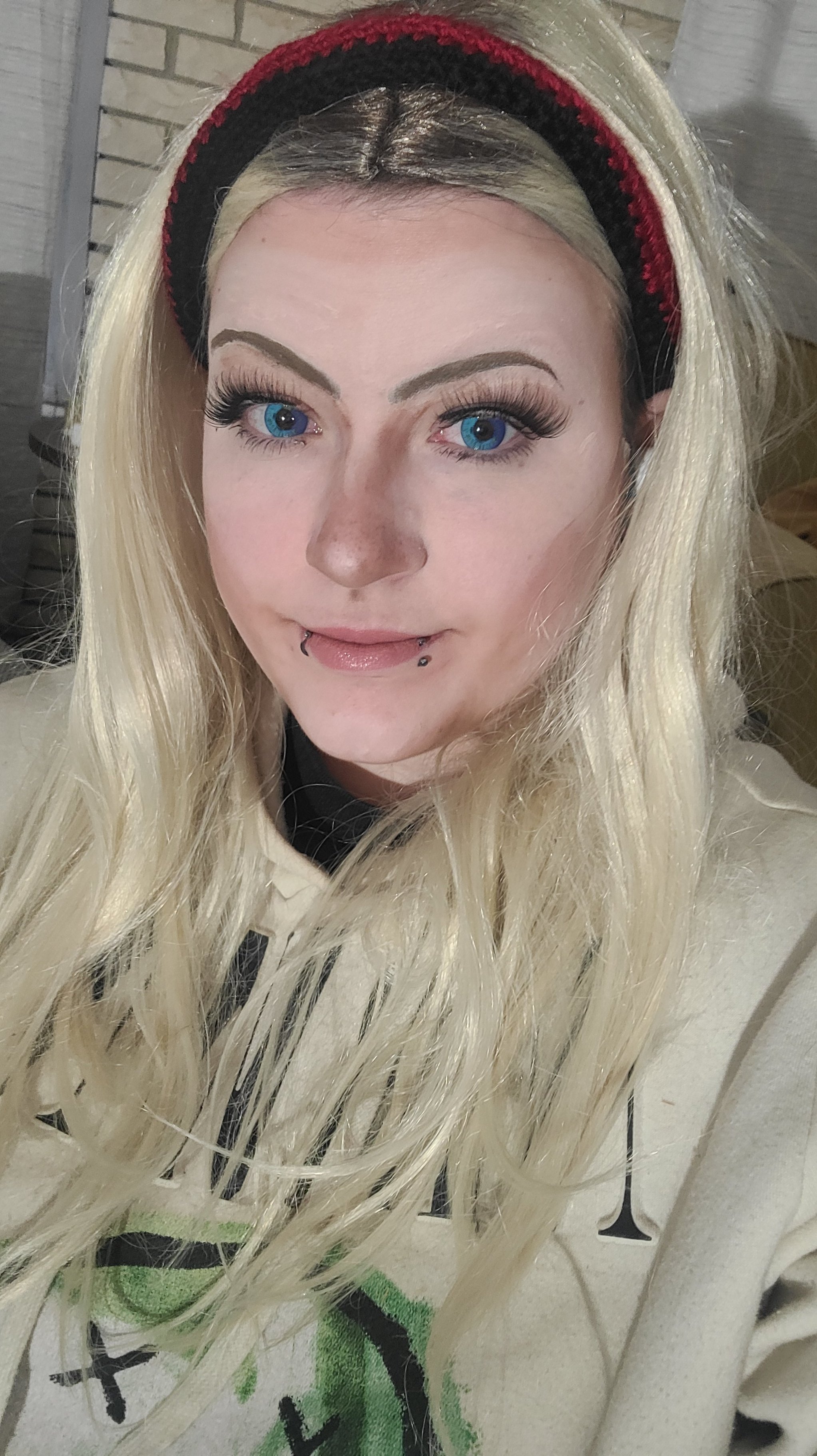 A young woman with long, wavy blonde hair wearing a red and black headband looks at the camera with bright blue eyes and has lip piercings.