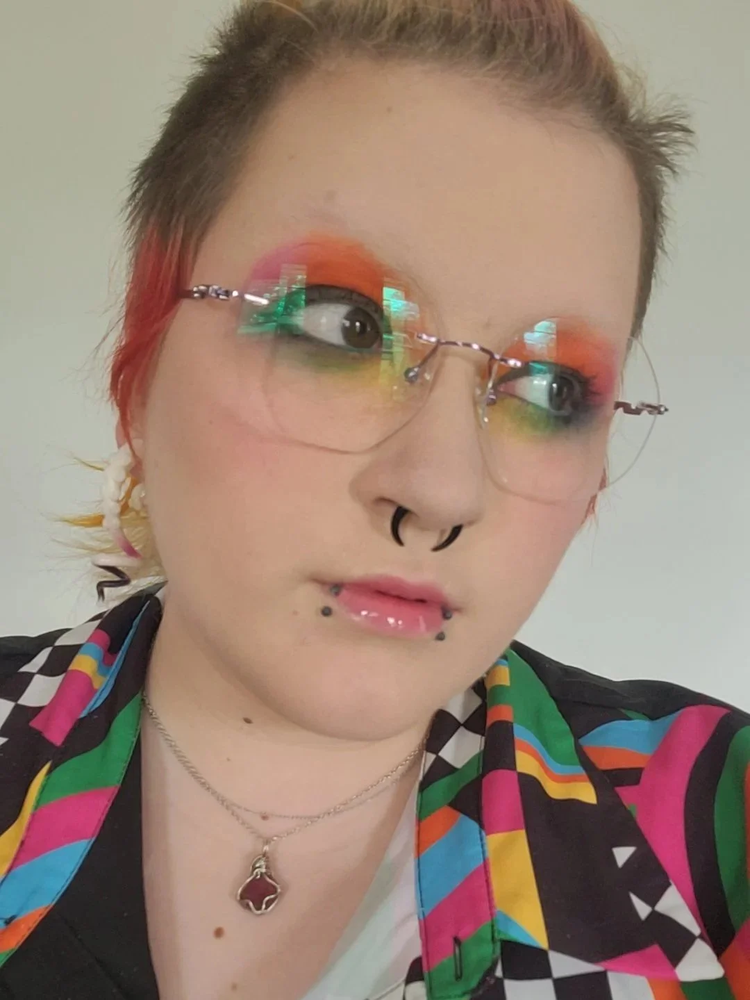 A person with rainbow-colored makeup, glasses reflecting colorful lights, multiple facial piercings, short hair with dyed red tips, wearing a vibrant patterned shirt and a black star pin, and a necklace with a heart-shaped pendant.