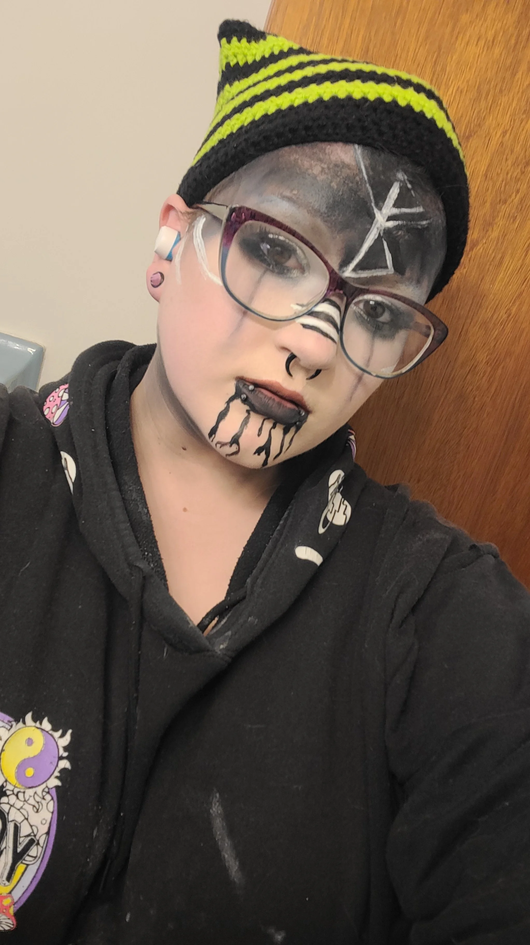 A person with face paint and virtual tattoos takes a selfie. They are wearing glasses, a black hoodie with a cartoon design, and a black and yellow striped knit cap.