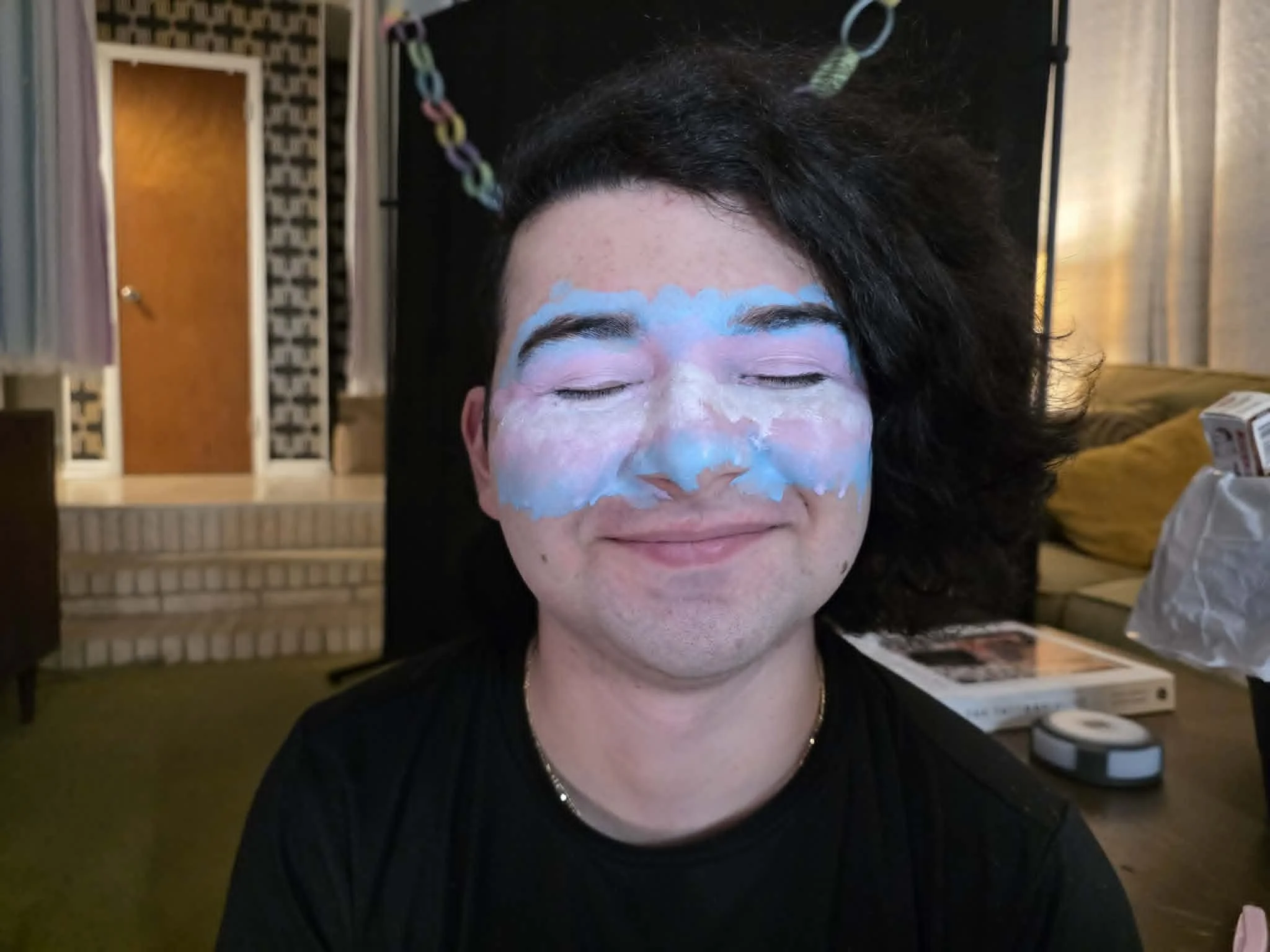 A person with makeup smeared across their face, eyes closed, in a room with curtains and shelves in the background.