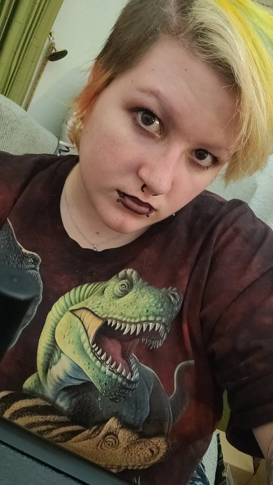 A person with short multicolored hair, dark makeup, and lip piercings wearing a graphic T-shirt with a dinosaur design taking a selfie indoors.