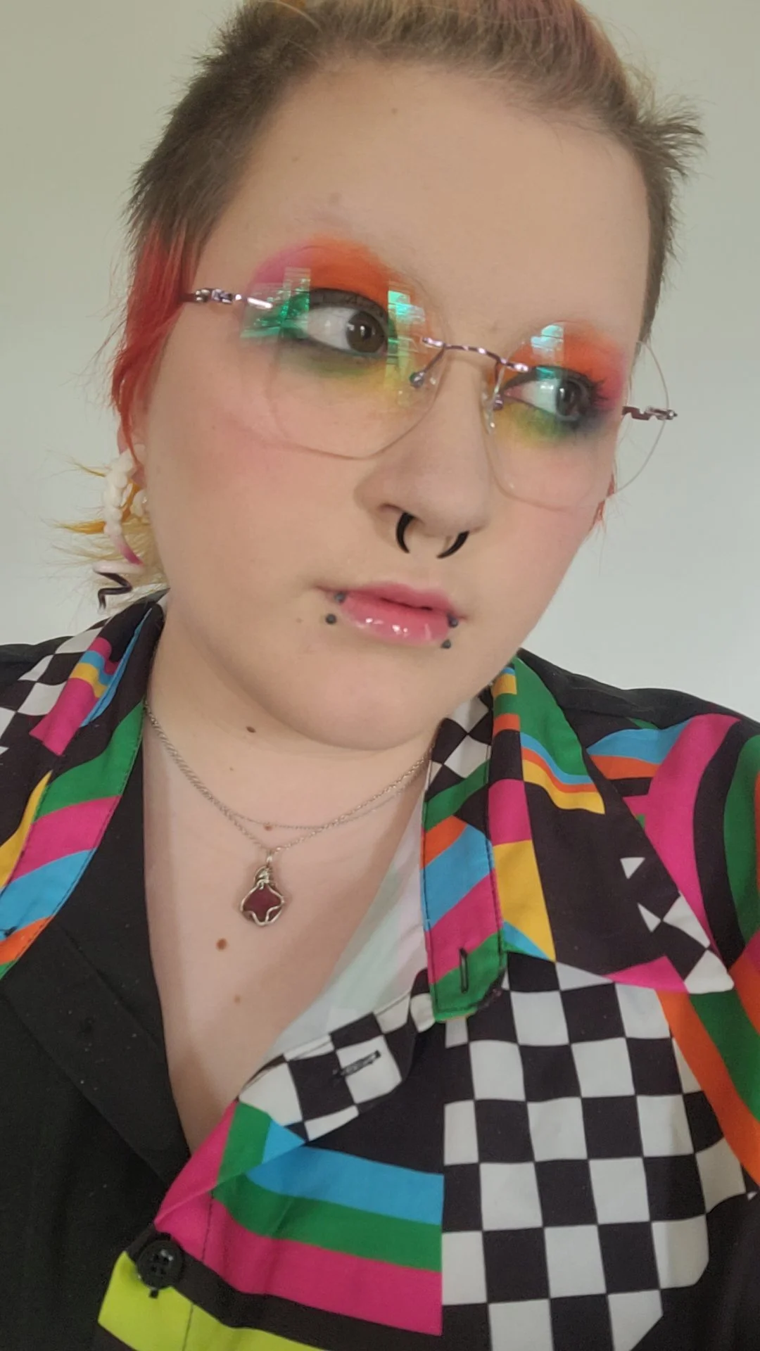 A person with short hair, wearing glasses with rainbow-colored lenses, colorful makeup with orange eyeshadow, and multiple facial piercings. They are dressed in a vibrant, patterned shirt and are wearing a silver necklace with a red gemstone pendant.