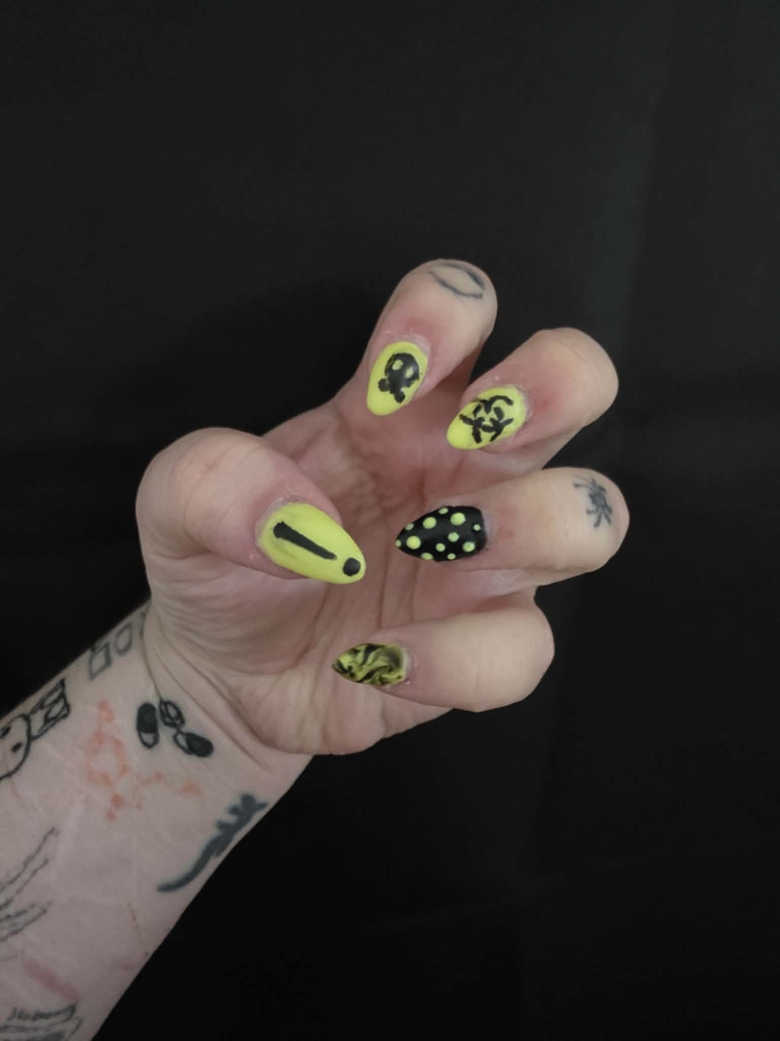 Close-up of a hand with yellow and black Halloween-themed nail art, including a ghost, a biohazard symbol, and polka dots, against a black background.
