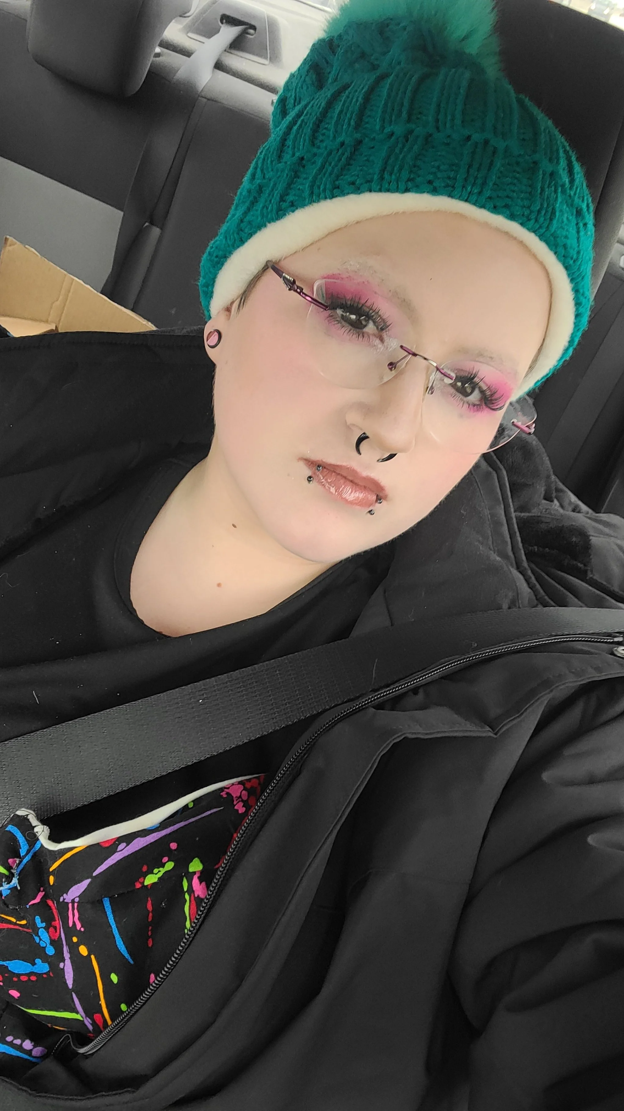 A person wearing a green knit hat with a pom-pom and pink eye makeup, glasses, and multiple facial piercings, sitting in a car seat with a black jacket and a black shirt with neon-colored splatters.