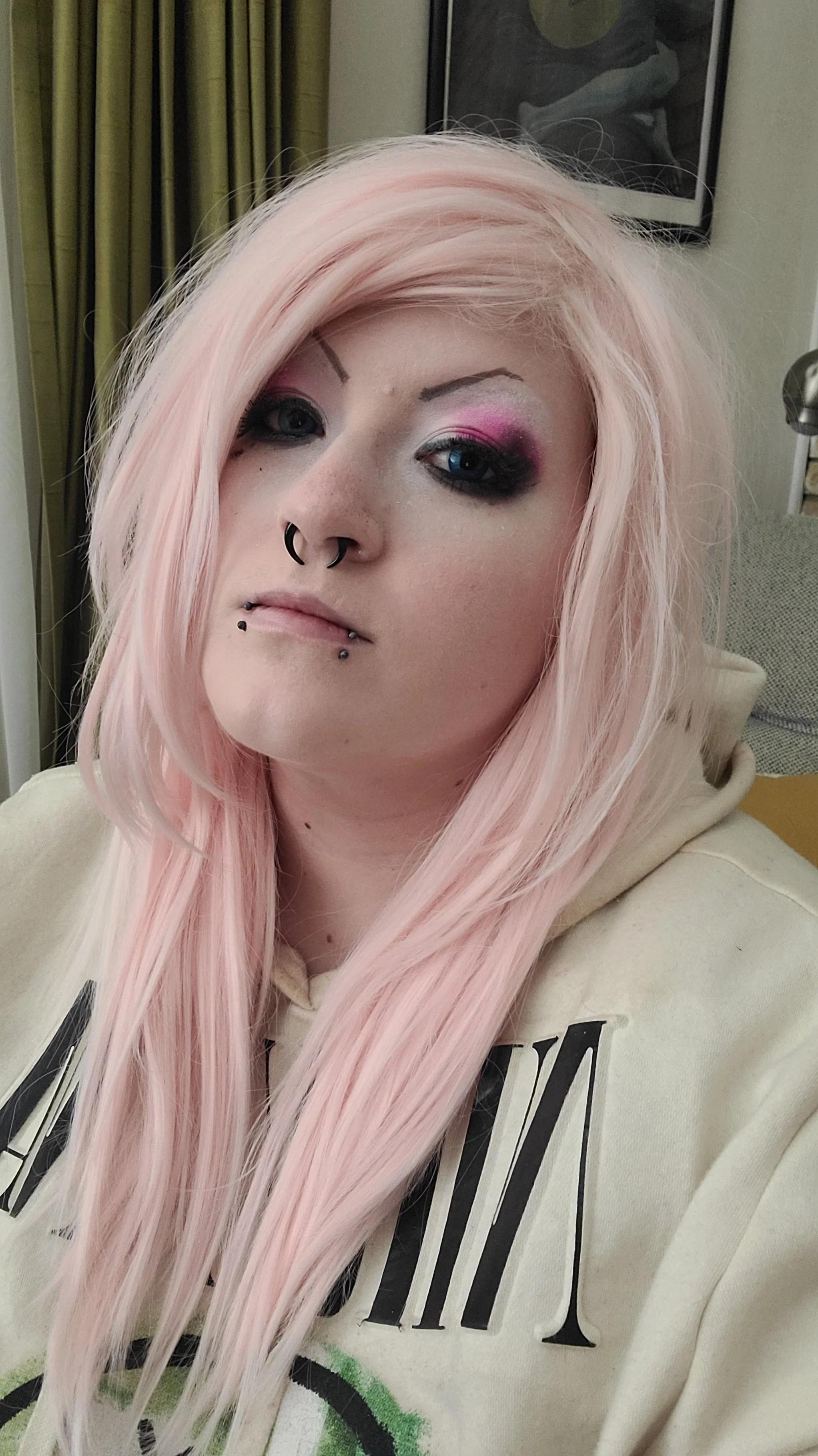 A woman with long pink hair, heavily applied eye makeup, and facial piercings, wearing a beige hoodie with black and green writing.