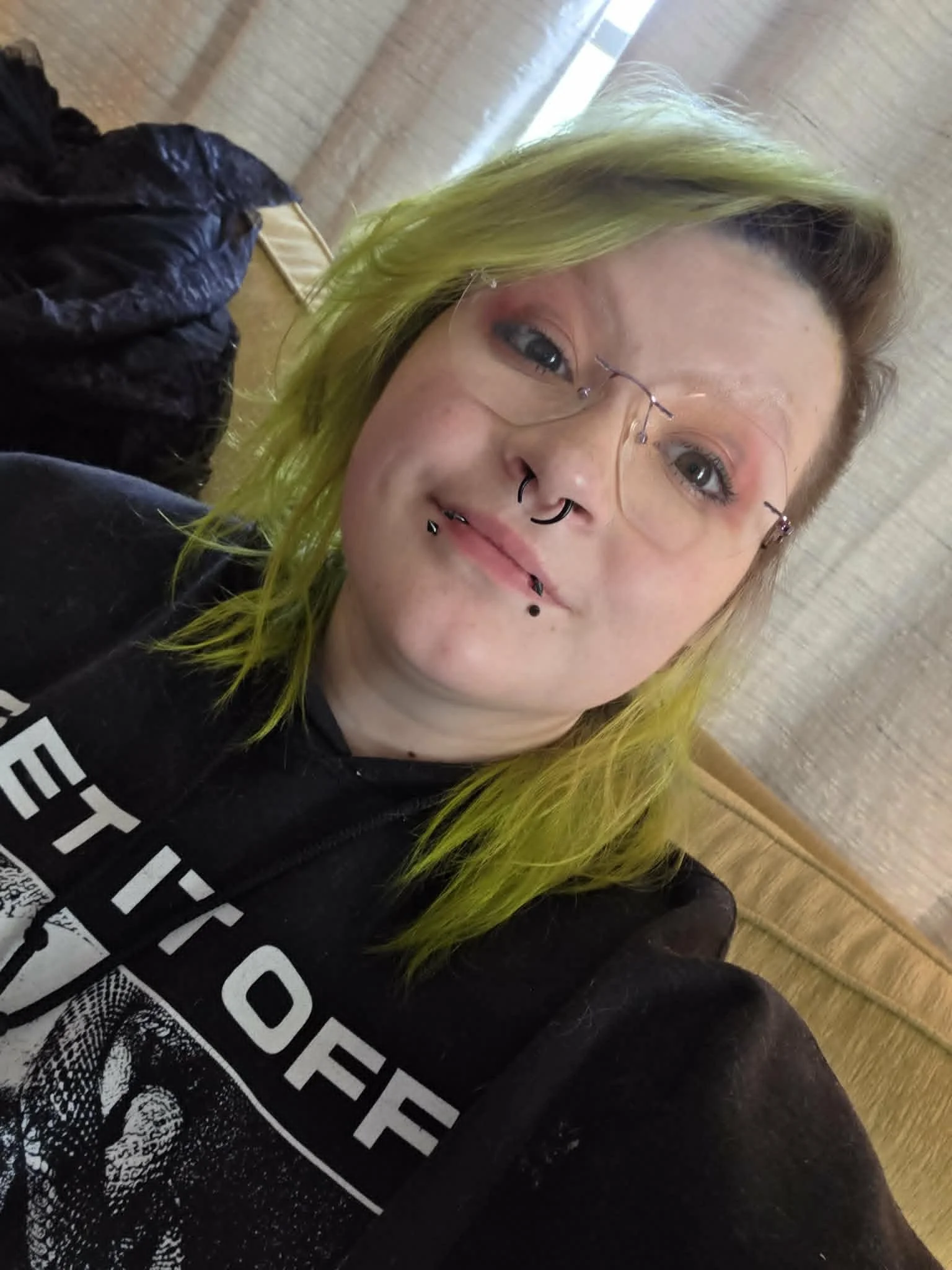 Close-up of a woman with green hair, wearing glasses and multiple facial piercings, smiling slightly in front of a wooden background.