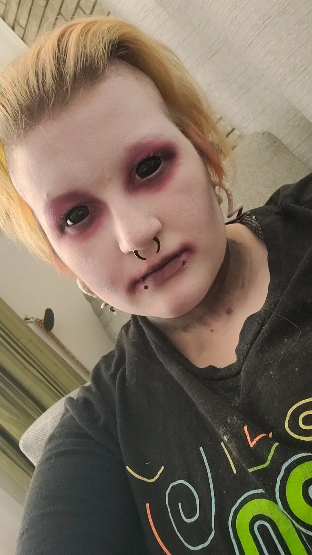 A person with pale skin, orange and blonde hair, and dark makeup around the eyes. They have multiple facial piercings, including a septum ring, lip piercings, and earrings. They are wearing a black shirt with a colorful logo or design.