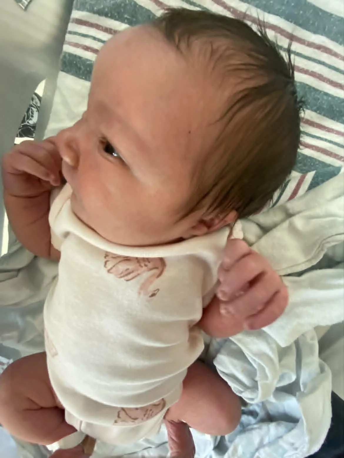 Close-up of a newborn baby with dark hair, lying on a hospital bed, wearing a cream-colored onesie with a flamingo print.