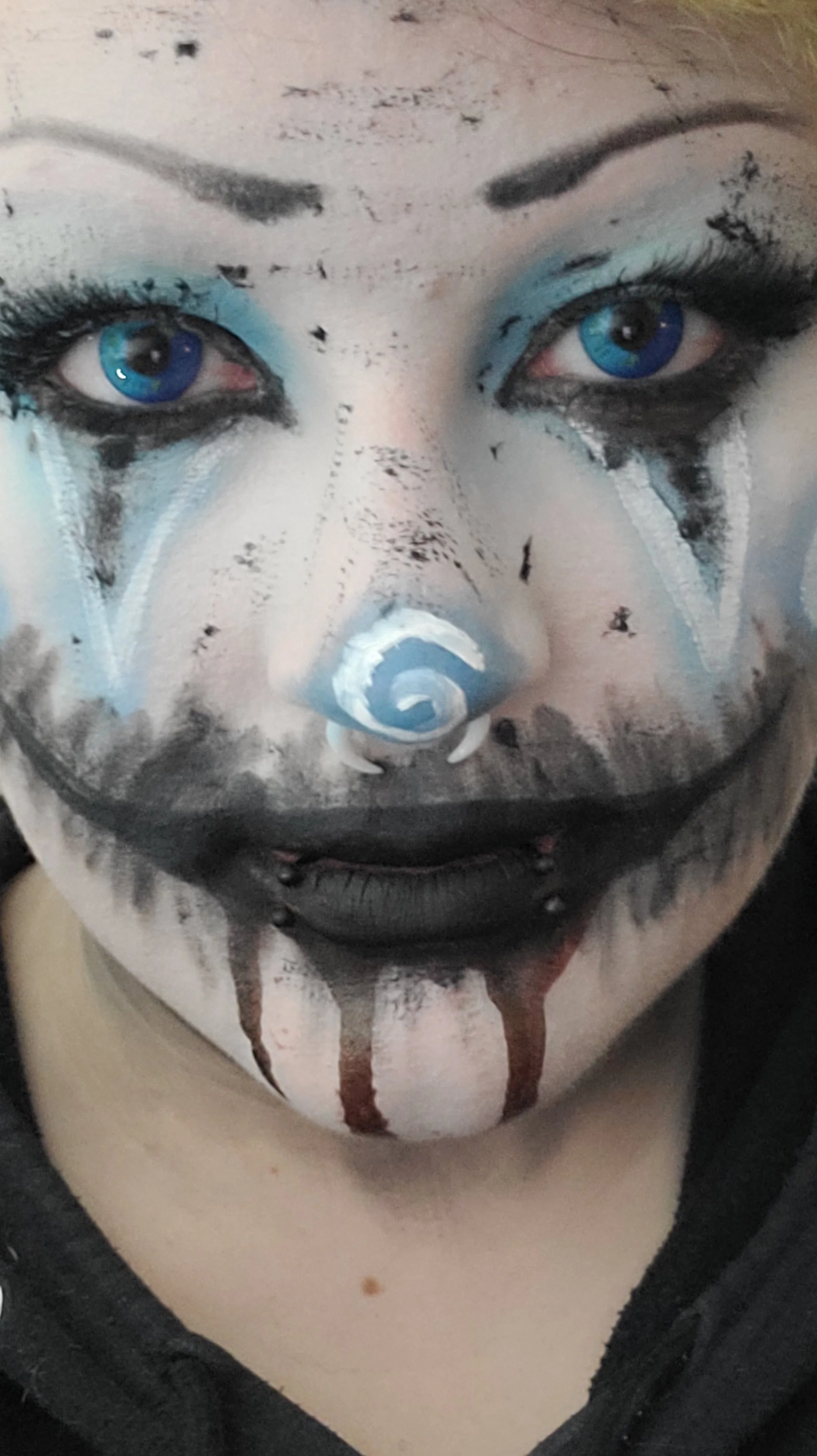 Person with clown makeup, featuring blue eye contacts, black and white face paint, and a nose painted with a spiral pattern, wearing a black shirt.