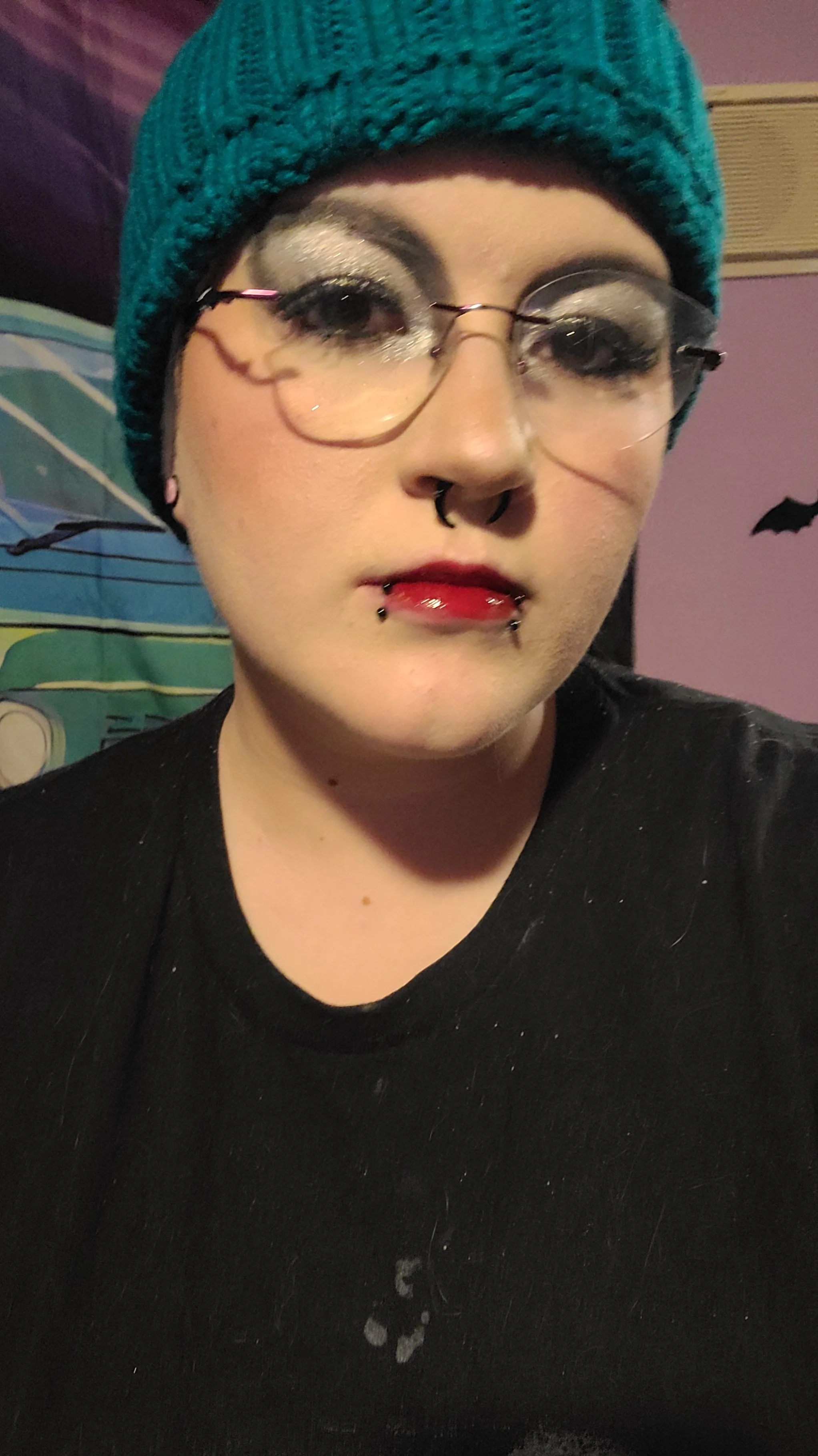 A person wearing glasses, a teal beanie, and makeup with dark lipstick, in a black shirt, with a purple wall and bat decoration in the background.