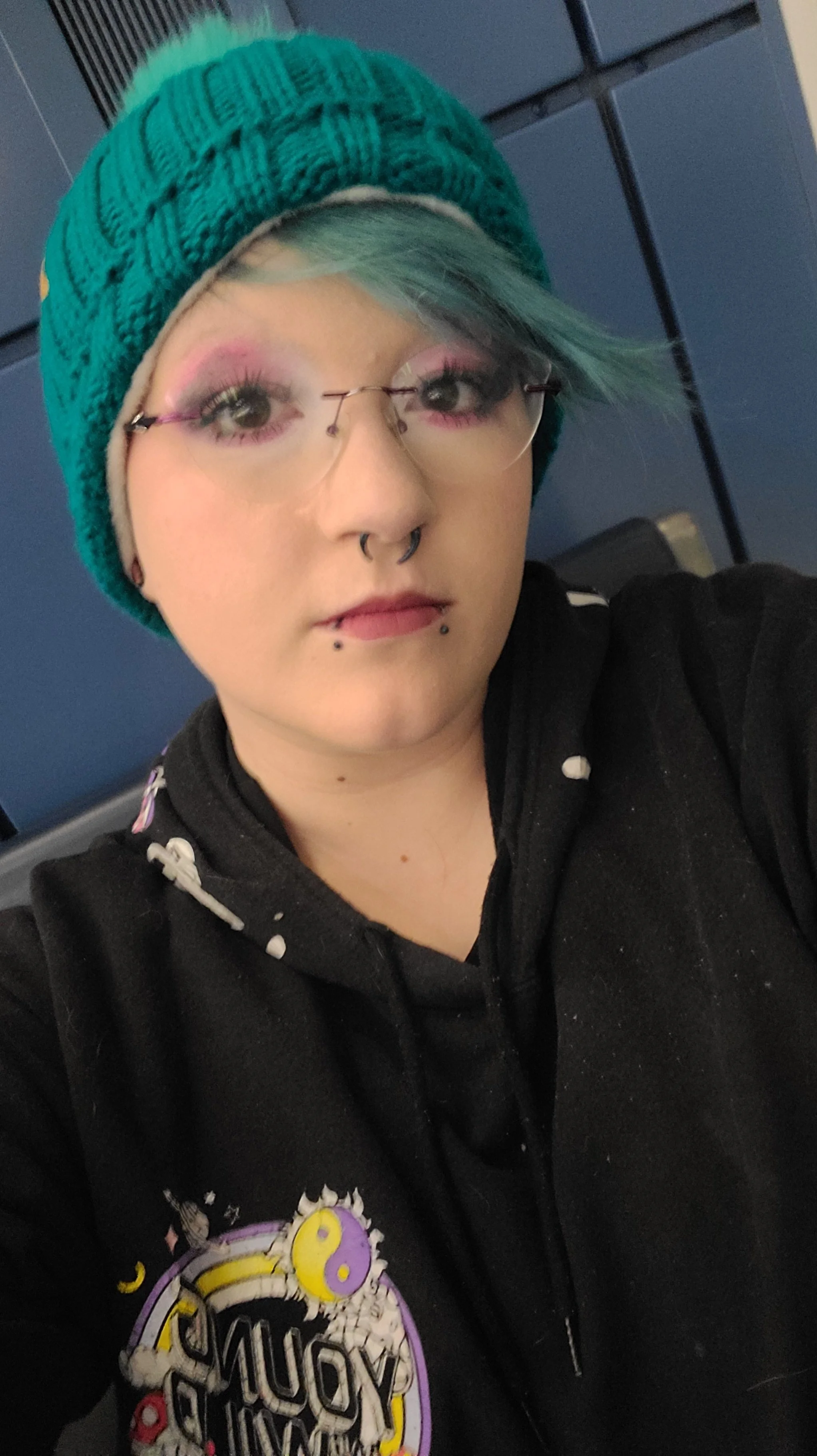A person with teal hair, wearing a teal knitted beanie, clear glasses, and a black hoodie with a colorful graphic design, taking a selfie in front of blue lockers.