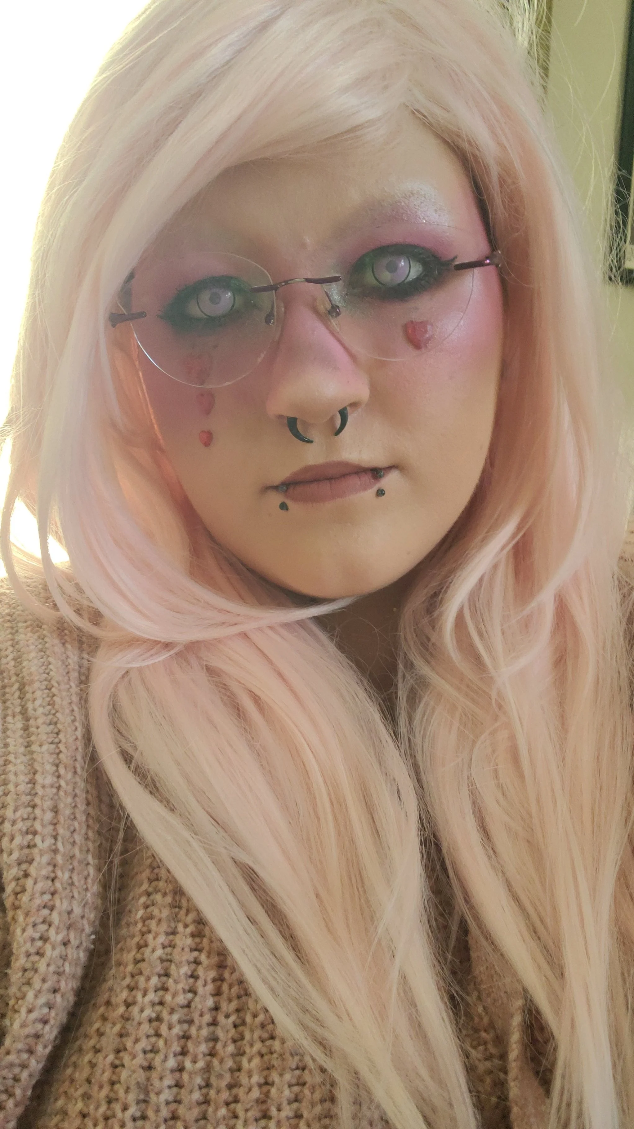 A young woman with long, wavy, platinum blonde hair and light pink skin. She has multiple facial piercings, including a septum and multiple lip piercings. She wears pink eyeshadow and glasses. She is wearing a beige, knitted sweater.