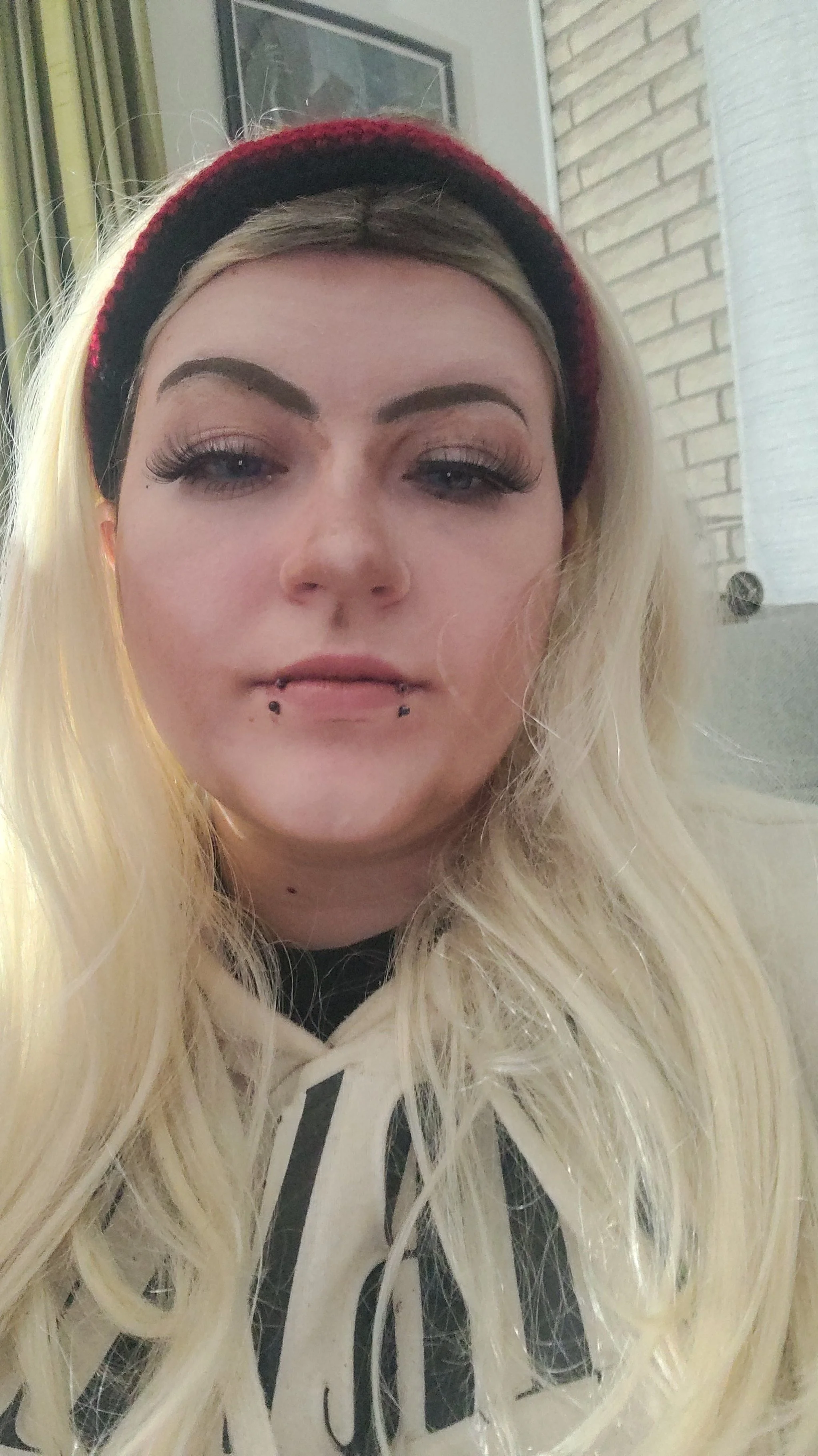 A close-up of a young woman with long platinum blonde hair, wearing a red and black headband and a white hoodie with black text.