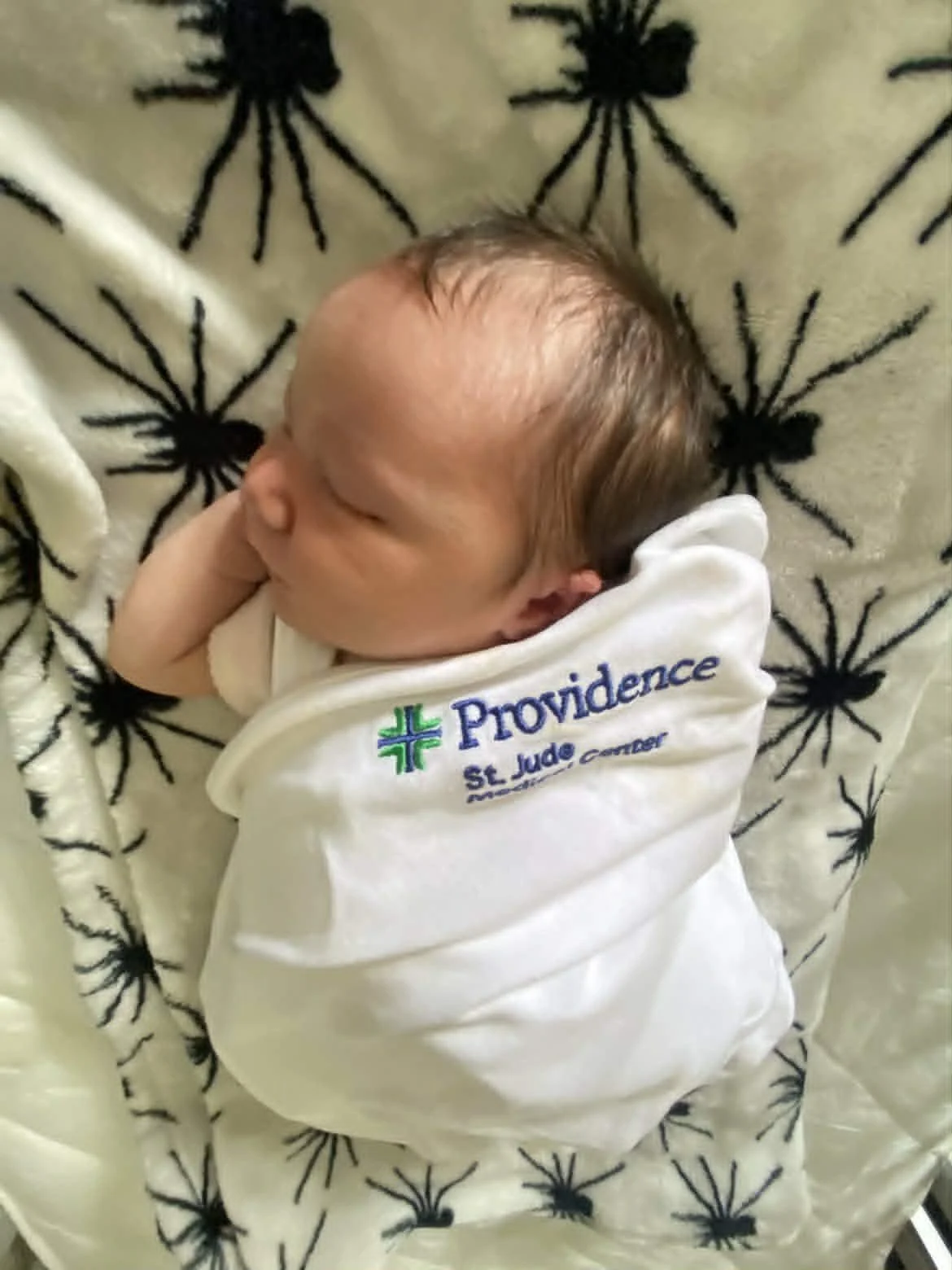 Newborn baby sleeping on a patterned blanket, wearing a white onesie with 'Providence St. Jude' logo.