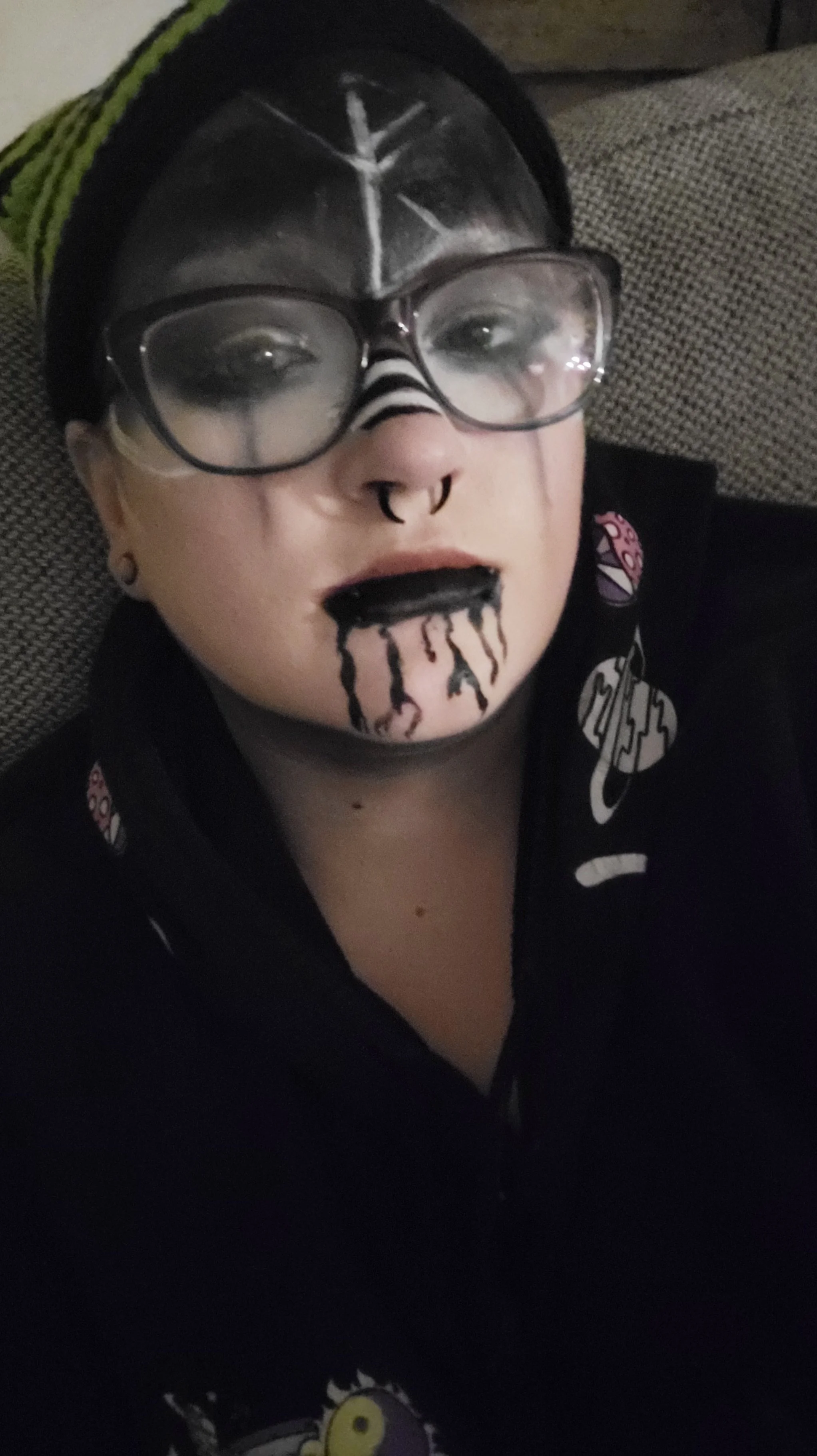 A person with black glasses, eyeliner, and dark lipstick with black drips, wearing a black shirt with colorful patches, sitting on a couch with a textured fabric background.